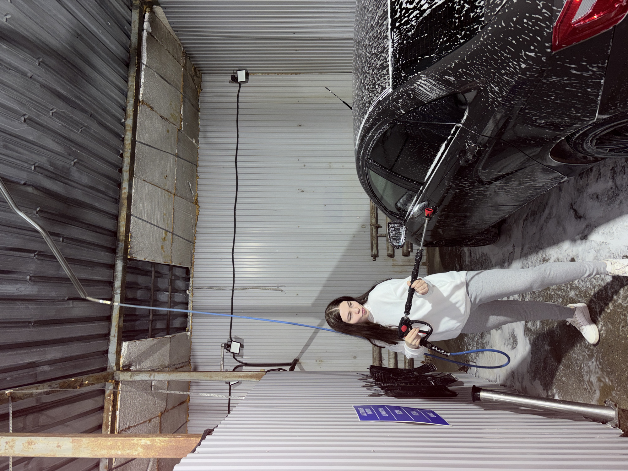 The image, taken in Chaikovsky, Russia, depicts a young woman engaged in washing a black car at a self-service car wash. She stands on the right side of the vehicle, holding a pressure washer with a black handle and red trigger, directing a stream of white foam onto the car's body. The black car is heavily covered in soap suds, with its red taillight and dark wheels visible.
The woman has long, dark hair and is casually dressed in a white long-sleeved sweatshirt, light grey sweatpants, and white sneakers. She appears focused on the task, with a calm expression and a faint smile.
The setting is an indoor car wash bay featuring corrugated metal walls, some sections appearing insulated or constructed from concrete blocks. Exposed metal beams and structural elements, displaying signs of rust in places, are visible above and around the washing area. The concrete floor is wet and covered in soap residue, reflecting the ongoing cleaning activity. Electrical fixtures and outlets are mounted on the walls.
In the foreground, a tall, shiny silver cylindrical post, likely part of the car wash equipment (e.g., vacuum or payment station), is visible. A blue sign with white text is affixed to the lower right wall, displaying service options such as "ВОДА" (Water), "ПЕНА" (Foam, with "АКТИВНАЯ" - Active - above it), "ВОСК" (Wax), and "ОСМОС" (Osmosis, referring to purified water). The overall atmosphere is functional and well-lit, typical of an industrial car wash environment.