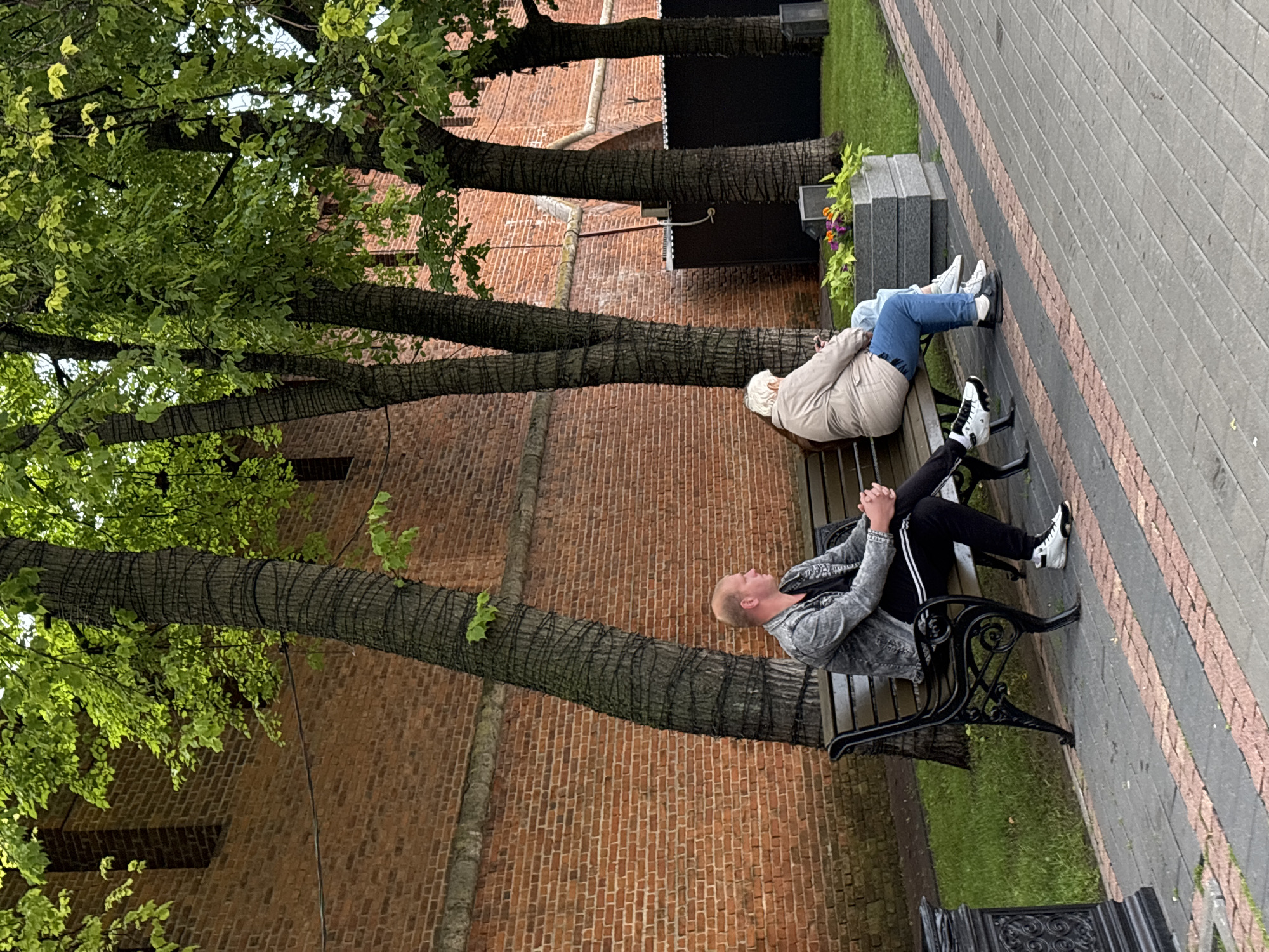 The image depicts an outdoor scene in Nizhny Novgorod, Russia, near the Kremlin walls. Two individuals are seated on a dark-colored bench. On the left, a man with a shaved head, wearing a denim jacket over a dark t-shirt and black pants with white stripes, sits with his hands clasped. On the right, an older person with light-colored hair, wearing a beige jacket and blue jeans, is seated and looking down at their phone.
Behind the bench, a tall brick wall, characteristic of the Kremlin, stretches across the background. Several trees with dark, textured bark and lush green foliage are positioned in front of the wall. The ground in front of the bench is a paved walkway with a pattern of gray and reddish-brown rectangular stones. To the right of the bench, a rectangular planter box filled with green foliage and some small purple and orange flowers is visible. The lighting suggests it is daytime, likely overcast given the diffused light.