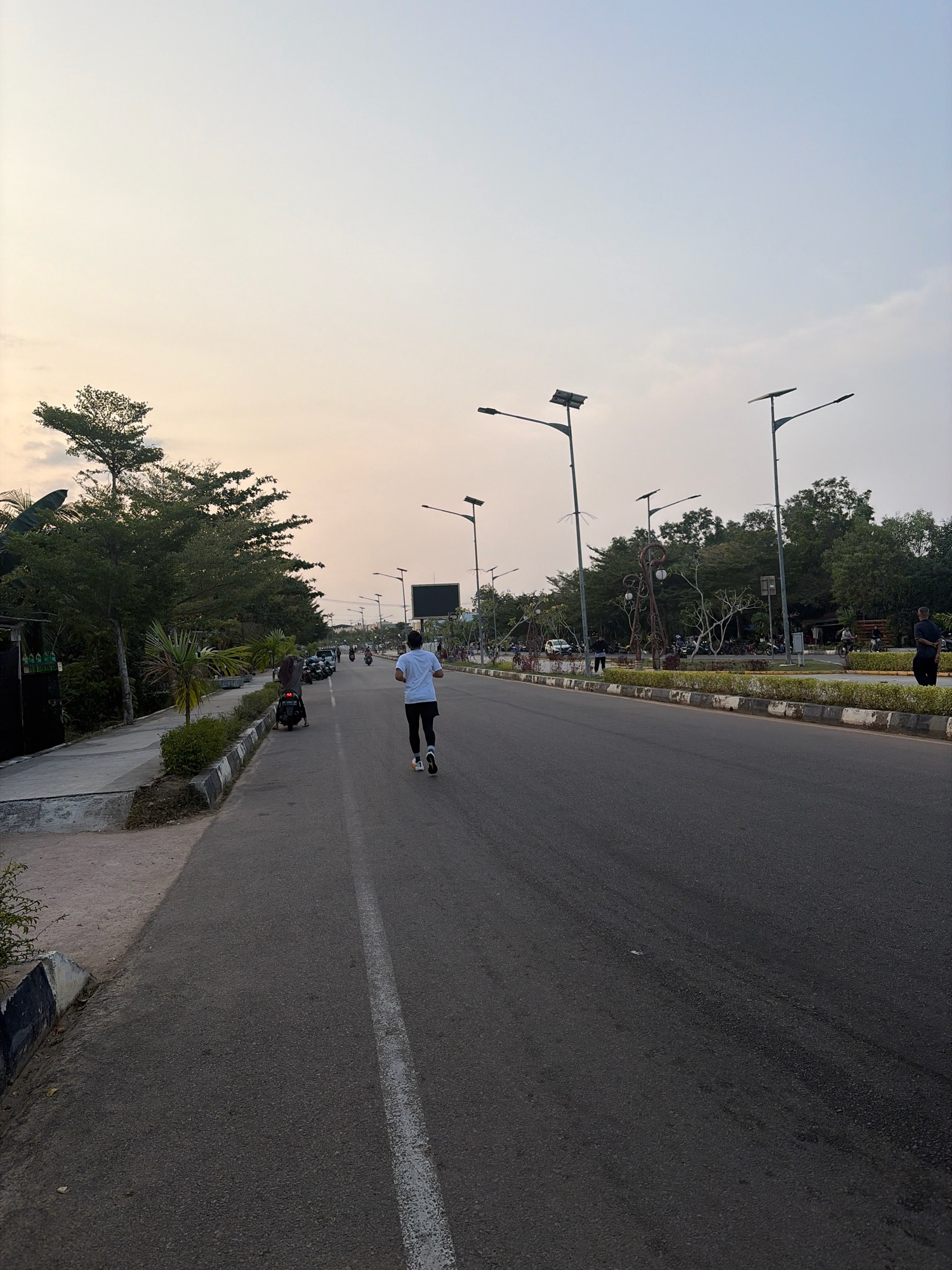 The image depicts a man running on a paved road in Tanjung Pinang, Indonesia, likely during the early morning or late afternoon due to the soft, diffused light. The road has a white line marking the center. On the left side of the road, there's a sidewalk with a low wall, followed by a line of trees and some buildings. Several motorcycles are parked along the roadside, and a few other people are visible in the background, some standing and others on motorcycles. Streetlights, some equipped with solar panels, line the right side of the road, which is bordered by a landscaped median strip with manicured bushes and decorative lampposts. The sky is a pale, hazy blue.