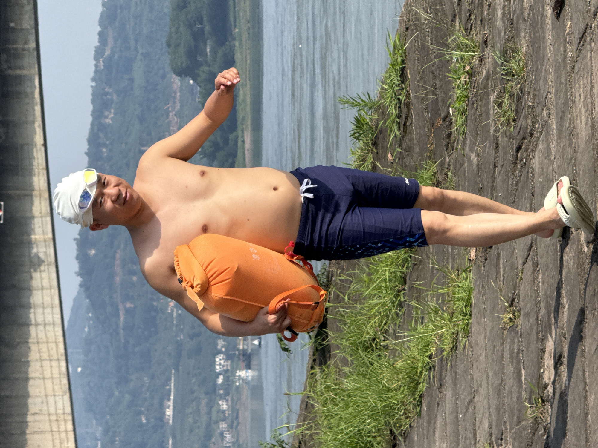 A man stands shirtless outdoors by a large body of water, likely a river or reservoir, in a bright, sunny daytime setting. He wears a white swimming cap, goggles, blue swim shorts, and light-colored sandals. He is holding an orange inflatable dry bag or swim buoy with both hands in front of him and looks towards the camera with a slight smile.
The man is positioned on a grey concrete or stone surface with green grass growing through its cracks, bordering the water. Across the calm water, distant mountains or hills are visible under a clear sky. To the far left, a substantial concrete wall or structure, possibly part of a dam or bridge, extends upwards. The man's attire and the swim buoy suggest he is either preparing for or has just completed a swim. Visible on the orange dry bag are some partially obscured white Chinese characters. The location context is Jiangjin, China.