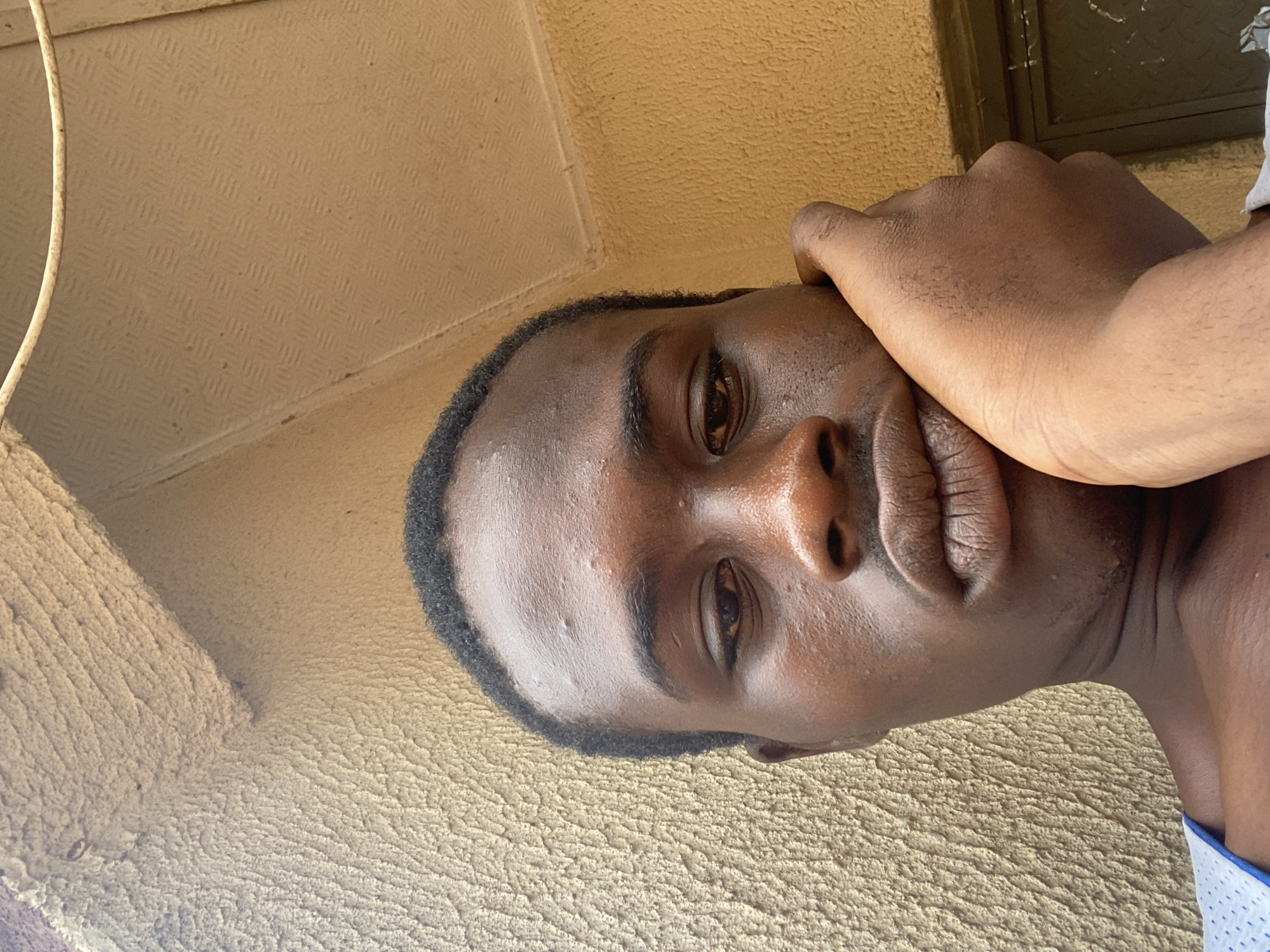 A close-up, low-angle, over-the-shoulder selfie shows a young Black male resting his right hand on his cheek, looking down with a pensive expression. He is outdoors, leaning against a textured, yellow-brown wall. Above him, a portion of a light brown, corrugated ceiling is visible, with a thin, rusty wire suspended across it. The man has short black hair and is wearing a white shirt with blue polka dots. The background consists of the textured wall and the corrugated ceiling, with a sliver of a dark-colored door or window frame visible in the lower right corner. The lighting suggests it is daytime.