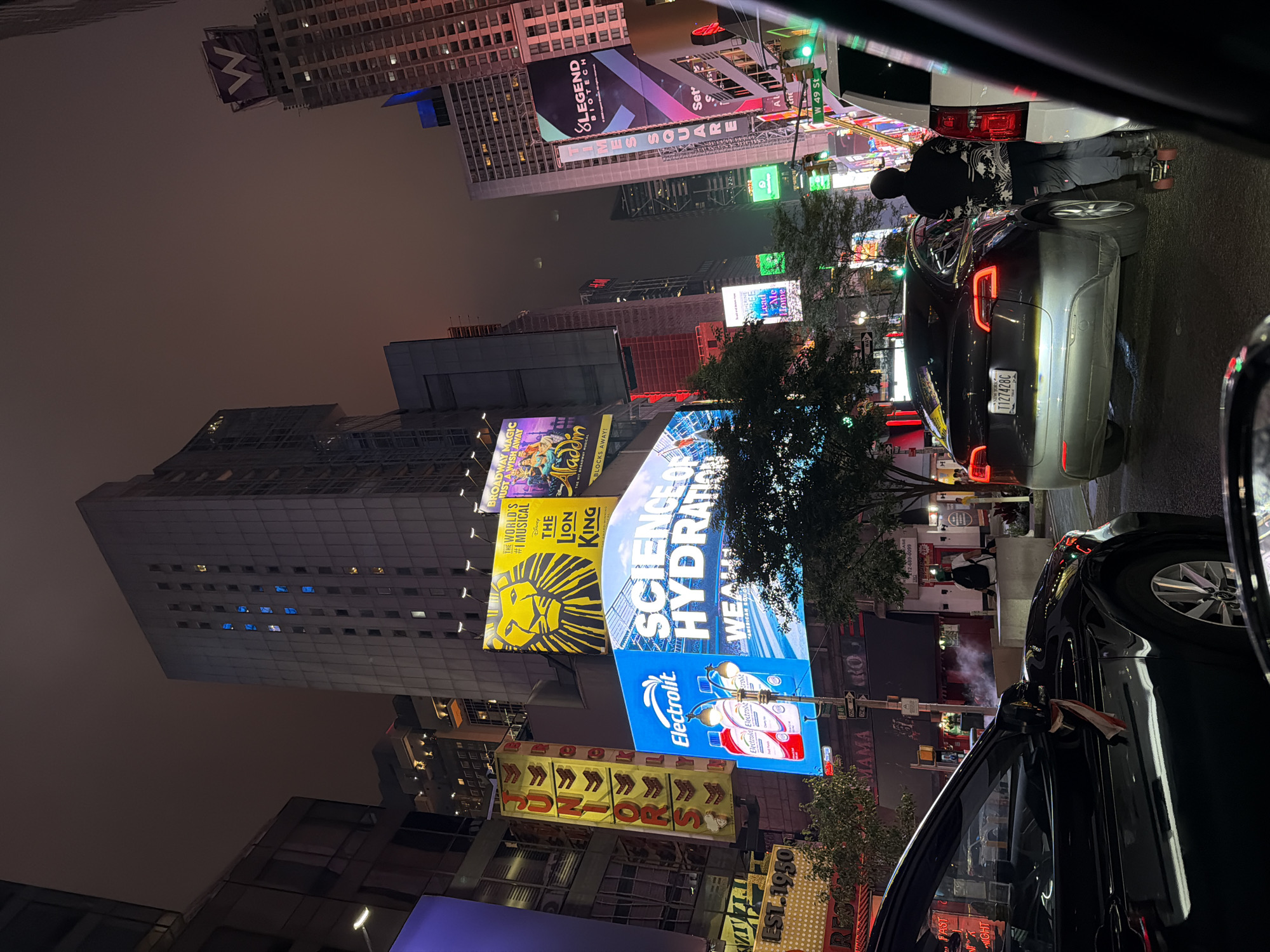 This image captures a nighttime view of Times Square in New York City, characterized by its iconic vibrant billboards and bustling atmosphere. The perspective is from inside a vehicle, looking out at the illuminated cityscape.
The main subjects include several large, brightly lit billboards advertising various products and shows. Prominently displayed are advertisements for "The Lion King" musical, "Electrolit" beverages, and a "Legend Biotech" billboard. The text "TIMES SQUARE" is also visible. The scene is illuminated by the artificial light of the billboards, creating a lively and energetic ambiance.
In the foreground, the car's interior is partially visible, with a black Tesla and a white SUV occupying the street. A person is seen standing near the rear of the white SUV, wearing casual clothing and possibly on roller skates. Steam or fog is emanating from somewhere in the scene, adding to the atmospheric quality.
The time of day is clearly night, indicated by the darkness of the sky and the brilliance of the illuminated signs. The weather appears to be slightly hazy or misty, contributing to the glow of the lights. The location is unmistakably Times Square, a well-known landmark in New York City, indicated by the street sign "W 49 St" and the density of advertising.
The overall impression is one of urban vibrancy and the distinctive energy of Times Square at night. The image captures the visual spectacle of advertisements and the flow of traffic in a famous metropolitan hub.