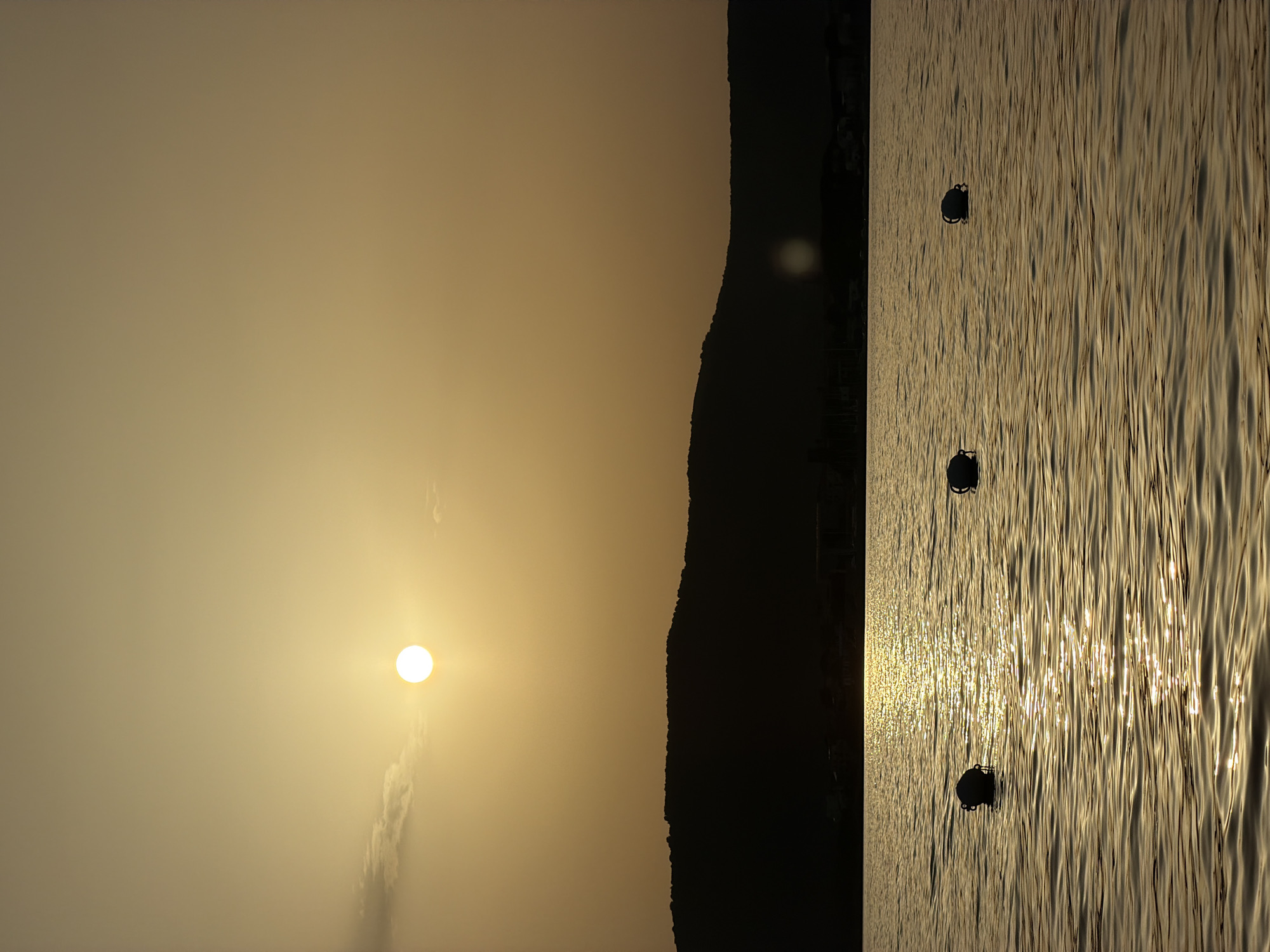 An atmospheric, low-angle shot captures a serene sunset over the waters of Alghero, Italy. The sun, a bright white orb, hangs in the hazy, golden sky, casting a shimmering reflection across the gentle waves. Silhouetted against the vibrant sky is a low, dark mountain range that forms the horizon. Along the water's surface, three round, dark buoys are visible, spaced evenly apart. The overall impression is one of tranquility and natural beauty.