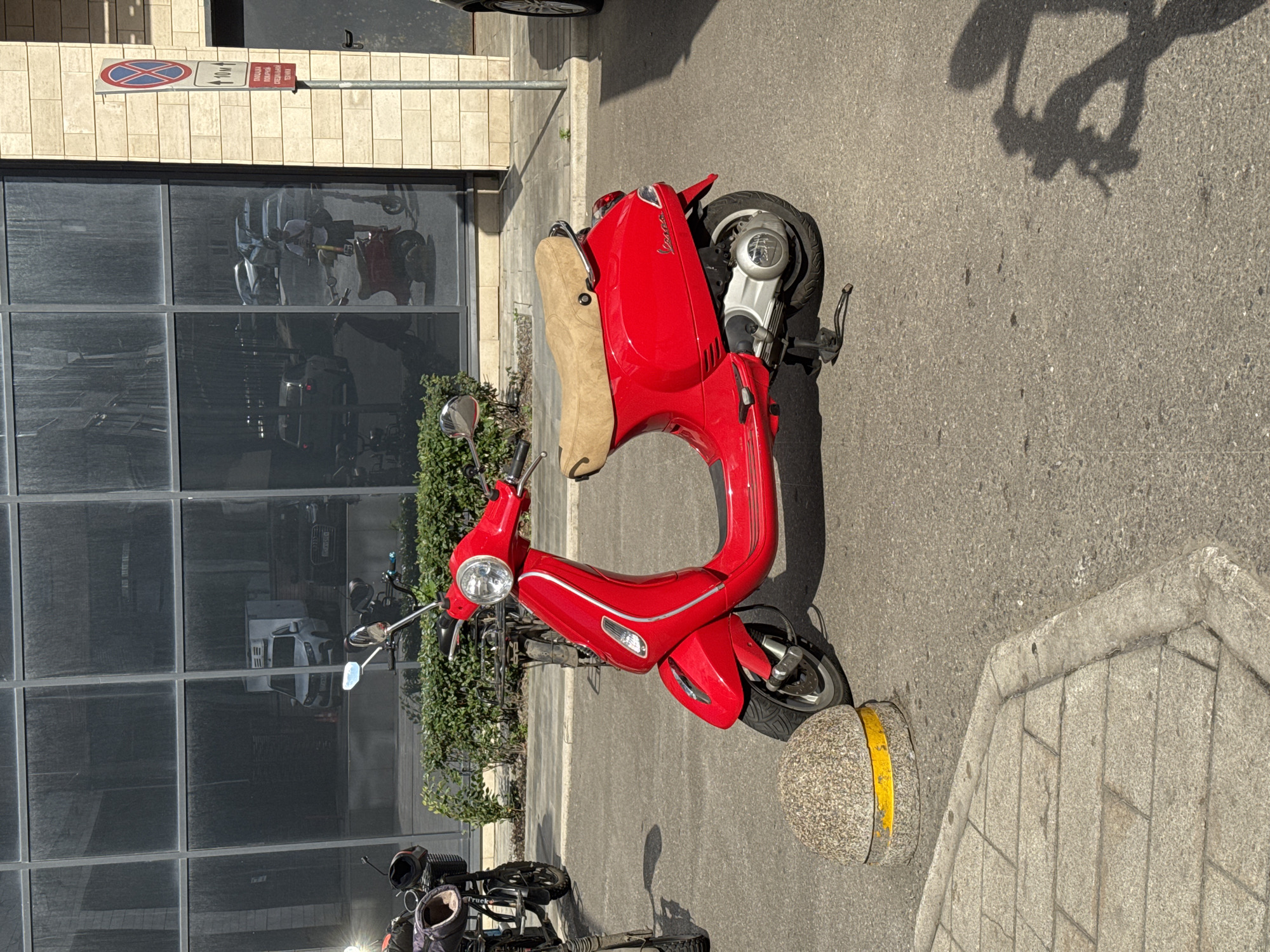 A bright red Vespa scooter is parked on a paved street next to a building with large, reflective windows. The scooter has a tan seat and chrome accents. A shadow of the scooter, elongated and distorted, is cast on the asphalt. In the reflection of the windows, other vehicles and possibly people can be seen. The building's facade is made of light-colored rectangular tiles. A traffic sign with a red circle and a diagonal line over a red "X," accompanied by a smaller sign indicating "10M" and some Cyrillic text, is mounted on a pole to the left of the scooter. The text on the sign appears to be in Russian and indicates a parking restriction. A round, textured bollard with a yellow stripe is visible near the scooter's rear wheel. The scene suggests a sunny day, indicated by the sharp shadow. The presence of the scooter and the building implies an urban setting, possibly in Moscow, Russia, as suggested by the Cyrillic text. The overall mood is neutral and observational.