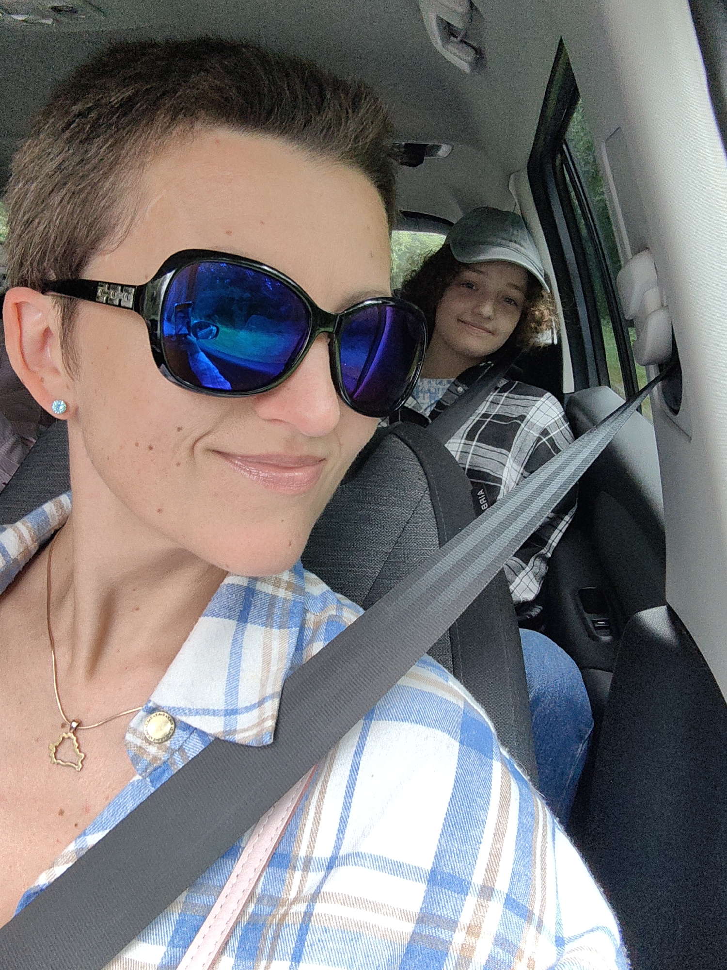 A selfie-style shot captures two individuals in the interior of a vehicle. The person in the foreground, on the left, is wearing large, dark sunglasses with blue reflective lenses and a blue and white plaid shirt. They have short, dark hair and are wearing a gold necklace with a pendant shaped like the outline of Belarus. A light blue stud earring is visible on their left ear. The seatbelt is prominently displayed across their chest.
In the background, on the right, a second person is seated, wearing a light-colored baseball cap and a black and white plaid shirt. They are looking towards the camera with a slight smile. The interior of the car is visible, including the seats, window, and parts of the car's roof. The scene appears to be outdoors, with greenery visible through the car window. The car is likely on a road within the city of Юнькаўскі сельскі Савет, Belarus, given the context of the request.