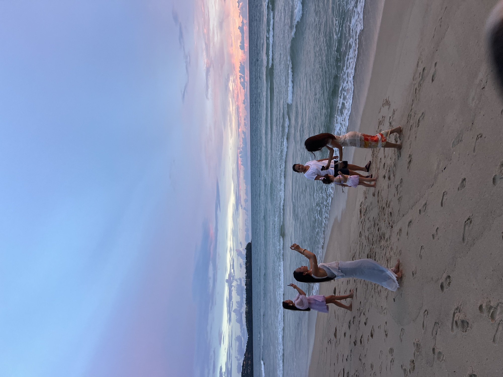 At dusk, a group of five people stand on a sandy beach near the ocean. The sky displays a gradient of soft blues, purples, and pinks, indicative of a sunset. Two women in the foreground on the left are holding up their phones, appearing to take pictures. A woman in a light blue dress is positioned in the center, also with a phone in hand. Further to the right, a woman in a patterned dress and a man in dark shorts and a white t-shirt are with a young child. The ocean has gentle waves reaching the shore, and footprints are visible in the sand. In the distance, a tree-covered landmass is visible on the horizon. The overall scene suggests a family or friends enjoying a beach outing during twilight.