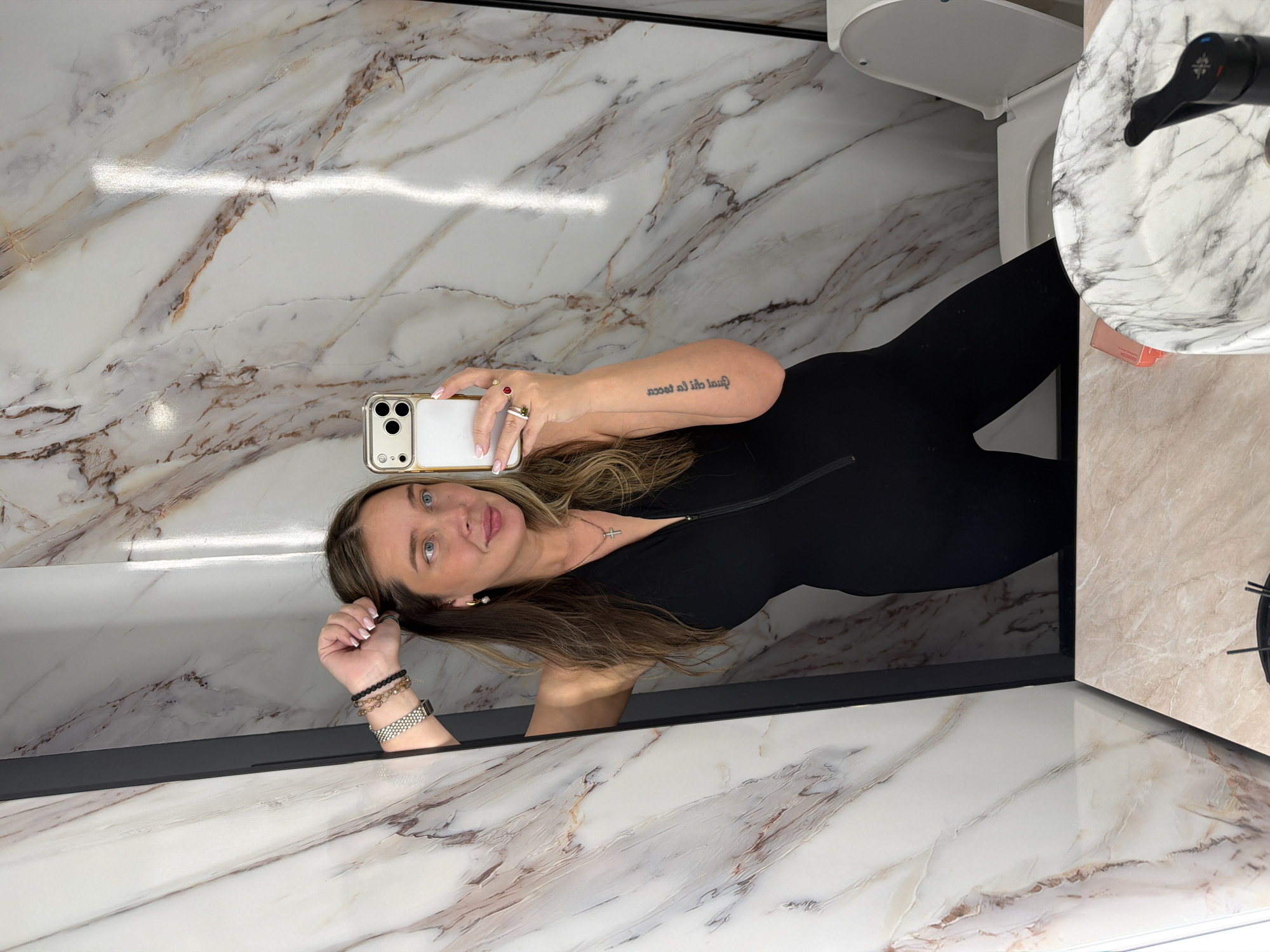 The image shows a woman taking a selfie in a bathroom. The woman is wearing a black outfit. She is holding a phone with a white back and a gold case. The bathroom has marble walls and countertops. There is a toilet and a sink in the background. The location cannot be determined with certainty as Moscow, Russia, based on visual information alone.