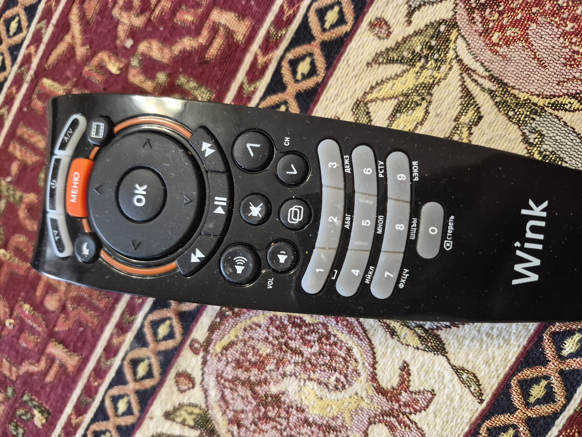 A black remote control, branded "Wink," is positioned diagonally across a richly patterned textile background. The remote appears well-used, with some dust visible on its surface. Its main features include a central circular directional pad with a prominent "OK" button. To the left of the directional pad is a bright orange "МЕНЮ" (Menu) button and an "A/V" input selector. Other control buttons include symbols for play/pause, rewind, fast forward, a film strip icon, "CH" (Channel) up/down, and "VOL" (Volume) up/down, along with a mute button.
On the right side of the remote, there is a numeric keypad with digits 0 through 9. Each number button also features Cyrillic letters beneath it, such as "АБВГ," "ДЕЖЗ," "ИЙКЛ," "МНОП," "РСТУ," "ФХЦЧ," "ШЩЪЫ," and "ЬЭЮЯ," used for text input. A "стереть" (delete) button is also visible near the "0" button. The brand name "Wink" is printed in white on the lower right side of the remote.
The background is a woven fabric, likely a rug or tapestry, displaying an intricate pattern in shades of maroon, cream, gold, and blue. The design features geometric shapes interspersed with stylized floral elements. No people or specific activities are depicted. The Cyrillic text on the remote and the "Wink" brand, associated with digital TV services in post-Soviet countries, provide a contextual link to the specified location of Yerevan, Armenia.