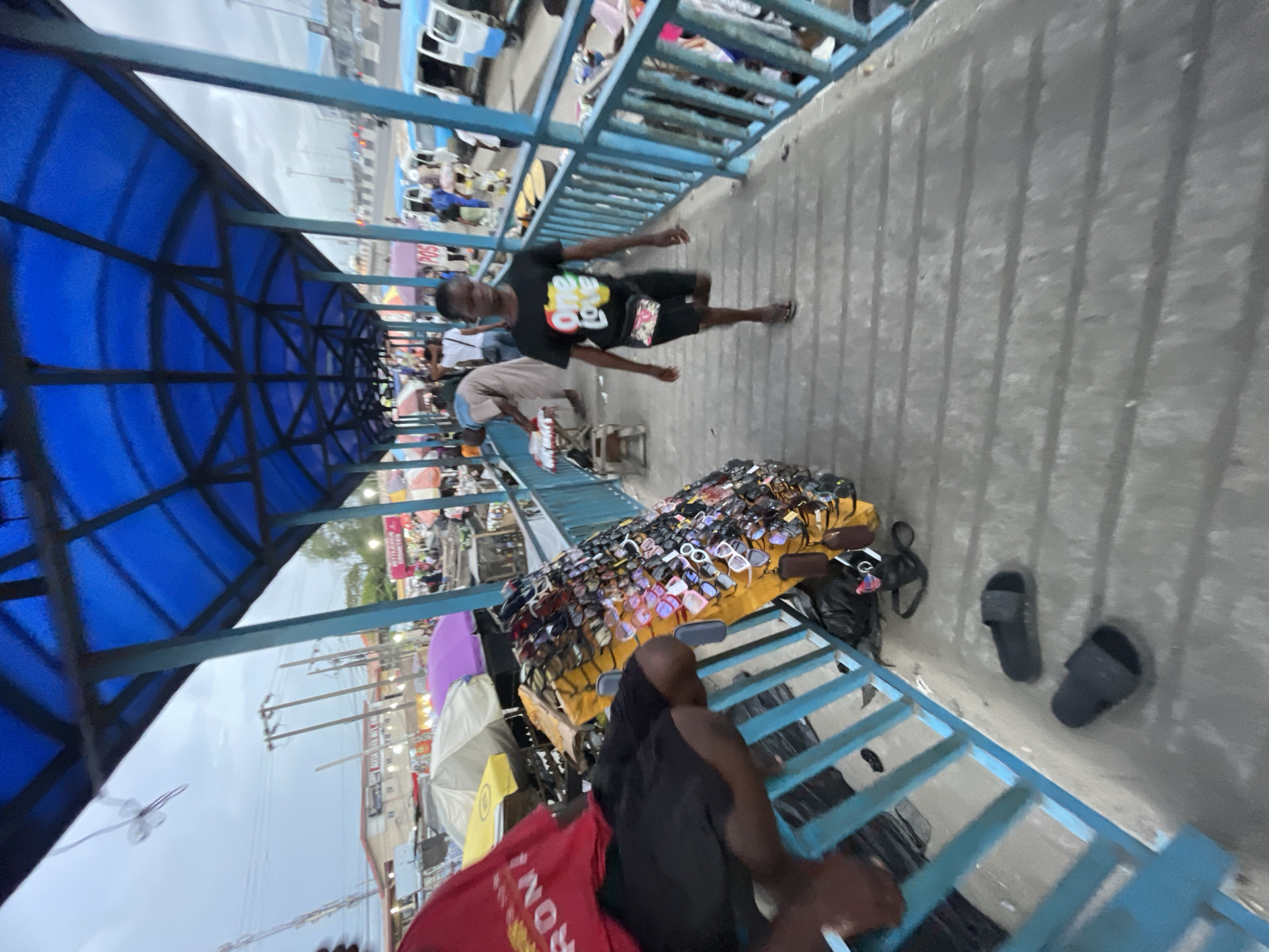 This photograph captures a bustling market scene in Lekki, Nigeria, likely taken from an elevated walkway or overpass. The image is oriented with the ground on the right.
The primary subjects are people engaged in commerce and movement. In the midground, a man with dark skin, wearing a black t-shirt with colorful "One Love" text and black shorts, walks along the paved walkway. He carries a small fanny pack across his chest. Further down the walkway, another person in lighter attire is visible near what appears to be another vendor. In the foreground, a stall packed with numerous sunglasses of various styles and colors is prominently displayed. Beside the stall, two black slip-on sandals rest on the ground. A person, partially obscured by the railing, is visible near the sunglasses display.
The setting is characterized by a blue-framed structure overhead, forming a translucent blue arched canopy providing shade to the walkway. A blue metal railing runs along one side of the elevated path. Below the walkway, a busy street or market area unfolds, with several vehicles (vans or small buses), numerous other stalls, and various market paraphernalia. Umbrellas of different colors are scattered amongst the stalls, indicating further vendor activity. Power lines and utility poles are visible against an overcast sky, suggesting it is daytime with cloudy weather. The scene conveys a sense of everyday market life and activity.