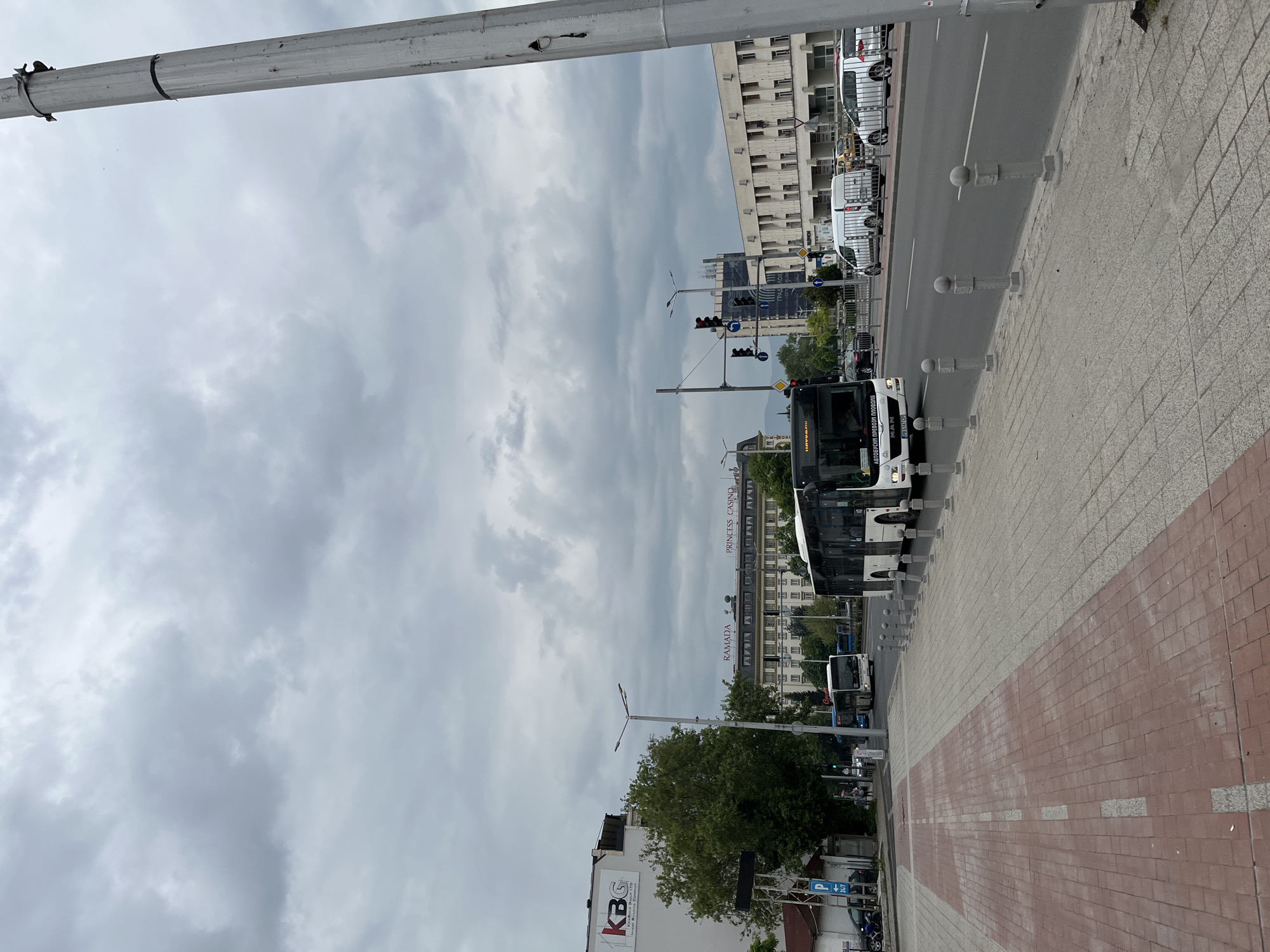 This photo shows a street scene in Plovdiv, Bulgaria. The sky is overcast. In the foreground is a gray and red brick sidewalk. To the right of the sidewalk is a series of gray bollards. A bus is partially visible, pulled over along the road. Cars are parked on the street further up. Buildings can be seen along the street, including a hotel labeled "Ramada" and "Princess Casino" further up the street. There are traffic lights in the distance. There is a tree in front of a white building with the red logo "KBG." There are motorcycles parked near the building.