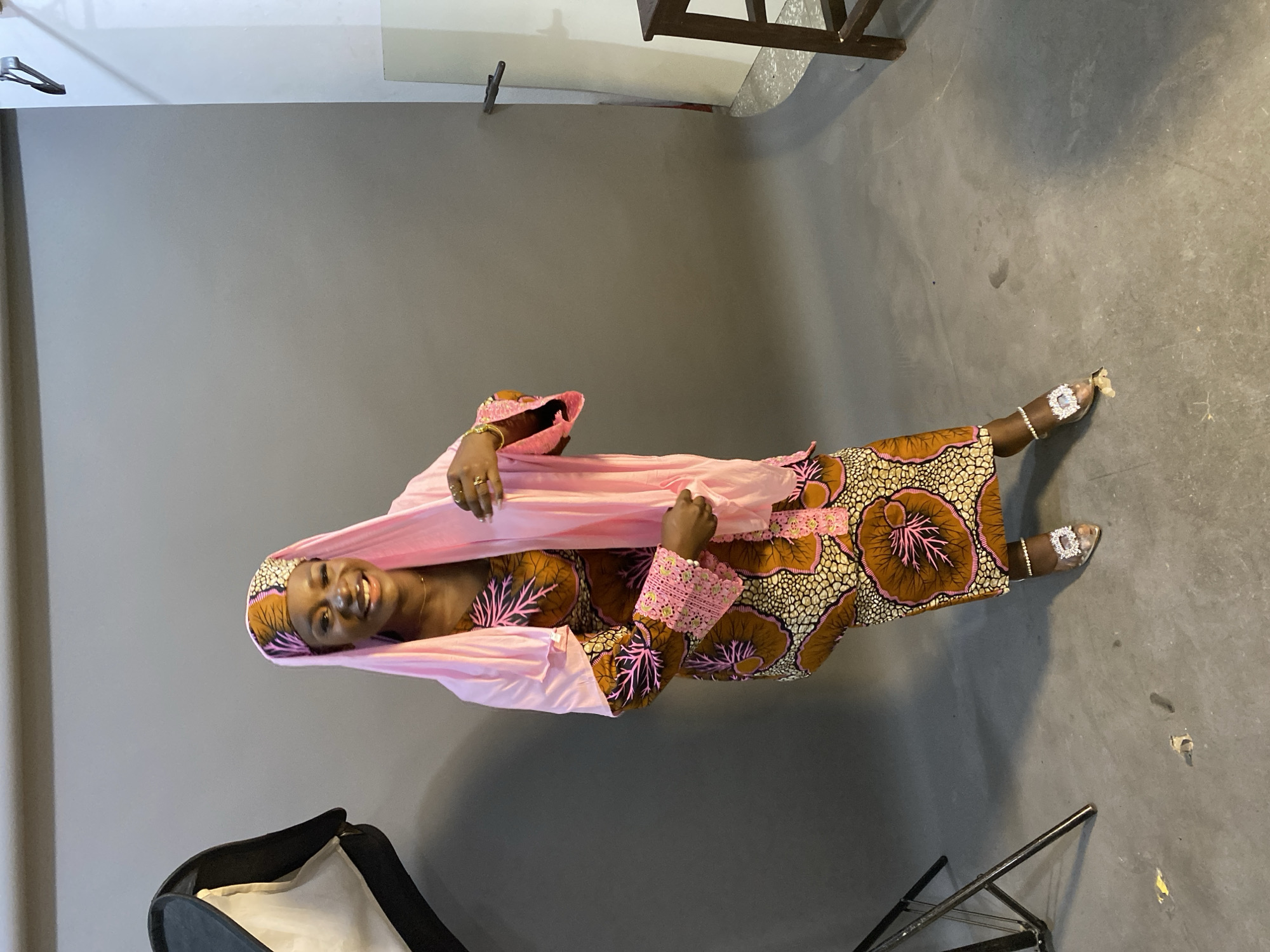 A joyful Black woman is captured in a vibrant portrait, standing upright and looking directly at the camera with a wide, genuine smile. She is dressed in a traditional long gown, likely an Ankara print, featuring a rich pattern of brown, pink, and cream/yellow hues with large, organic shapes. The dress has long sleeves adorned with intricate pink lace detailing at the cuffs. She elegantly drapes a plain pink headscarf or shawl over her head and shoulders.
Her hands, decorated with gold-colored rings and bracelets, gently hold the pink fabric. On her feet, she wears embellished open-toed sandals or heels that feature prominent, sparkling, possibly diamante, adornments.
The setting appears to be an indoor photography studio, characterized by a plain, solid grey wall as the background and a matching grey concrete floor. The upper portion of the background transitions to a lighter, possibly white, area, suggesting a ceiling or a different wall section. In the lower left corner, part of a black and white photography reflector or softbox stand is visible, and in the lower right, the metal frame of a folding chair or similar studio equipment can be seen. The floor shows some minor scuffs and marks.
The woman's bright smile conveys happiness and confidence. The traditional attire and vibrant colors suggest a cultural celebration or formal event in the context of Gura Top, Nigeria. There is no visible text in the image.