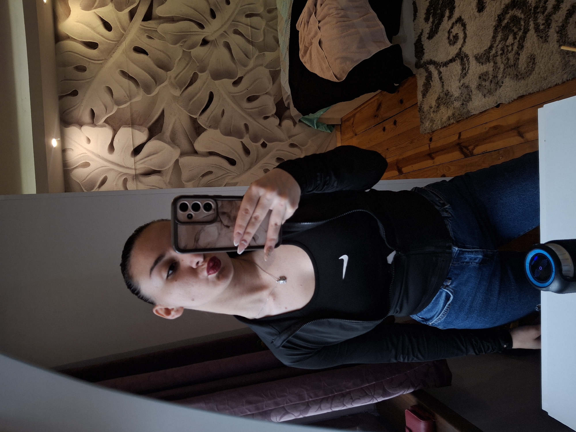 A person is taking a selfie in what appears to be a room in a house. The individual is wearing a black top with a white Nike logo and a black zip-up jacket, along with blue jeans. The background features a wall with a textured monstera leaf pattern, wooden floors covered with a rug, and a white table. The reflection in the mirror reveals a dark ceiling. No information in the photo confirms the location as Buhovo, Bulgaria.