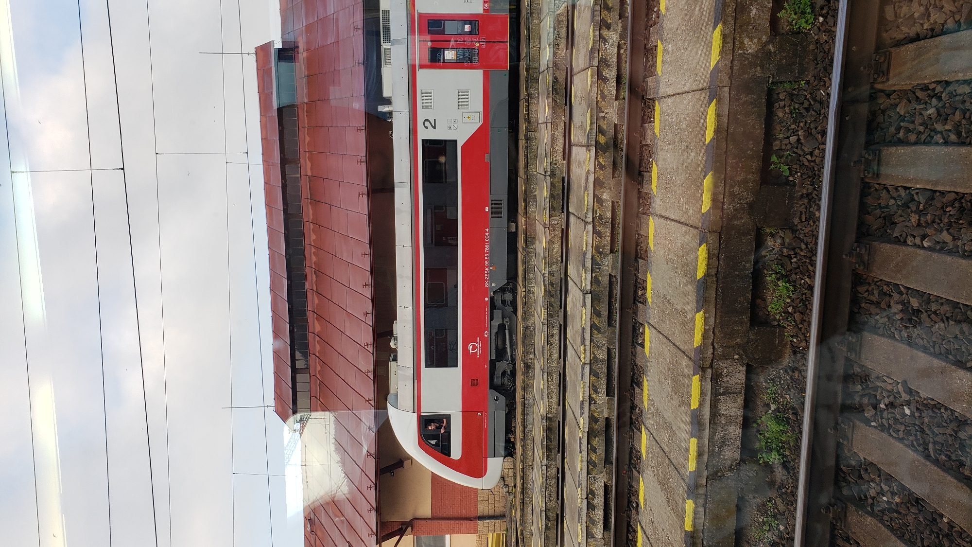The image shows a red and white train stopped at a station in Margecany, Slovakia. The train is a modern electric multiple unit, indicated by the number "2" on its side. A person is visible in the driver's cabin. The train is on a track with a concrete platform next to it, marked with yellow lines. In the background, a building with a red tiled roof is visible, which is part of the Margecany train station. Overhead power lines for the electric train are also present. The sky is overcast.