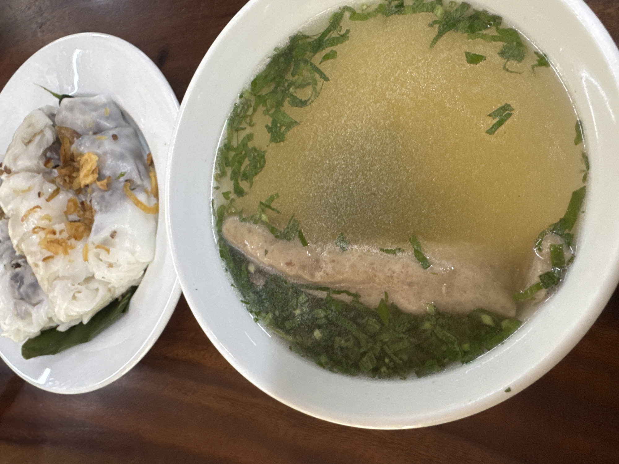 Two white plates of Vietnamese food are arranged on a brown wooden surface. The plate on the left contains several white rice noodle rolls topped with fried shallots, lying on a dark green leaf. The plate on the right is a bowl of clear broth soup garnished with chopped green herbs and featuring a pale, oblong piece of meat. The location is presumably a restaurant in the city of Hà Giang 1 Ward, Vietnam, based on the nature of the food.