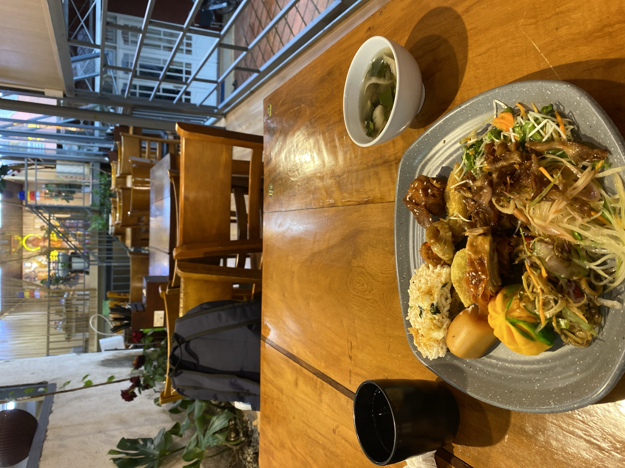 The image shows a close-up of a wooden table at what appears to be a restaurant in Da Lat, Vietnam. In the foreground, a grey plate is filled with a variety of food items, including rice, meat dishes, and a salad. To the left, a black cup holds a dark liquid. To the right, a white bowl contains a clear soup with vegetables. The table is set in a dining area with several wooden chairs and tables visible in the background, along with some greenery and architectural elements that suggest an indoor or semi-outdoor setting. The lighting suggests it is either evening or an overcast day. The background reveals more of the restaurant's interior, with decorative lighting and a glimpse of what might be a second floor or balcony.