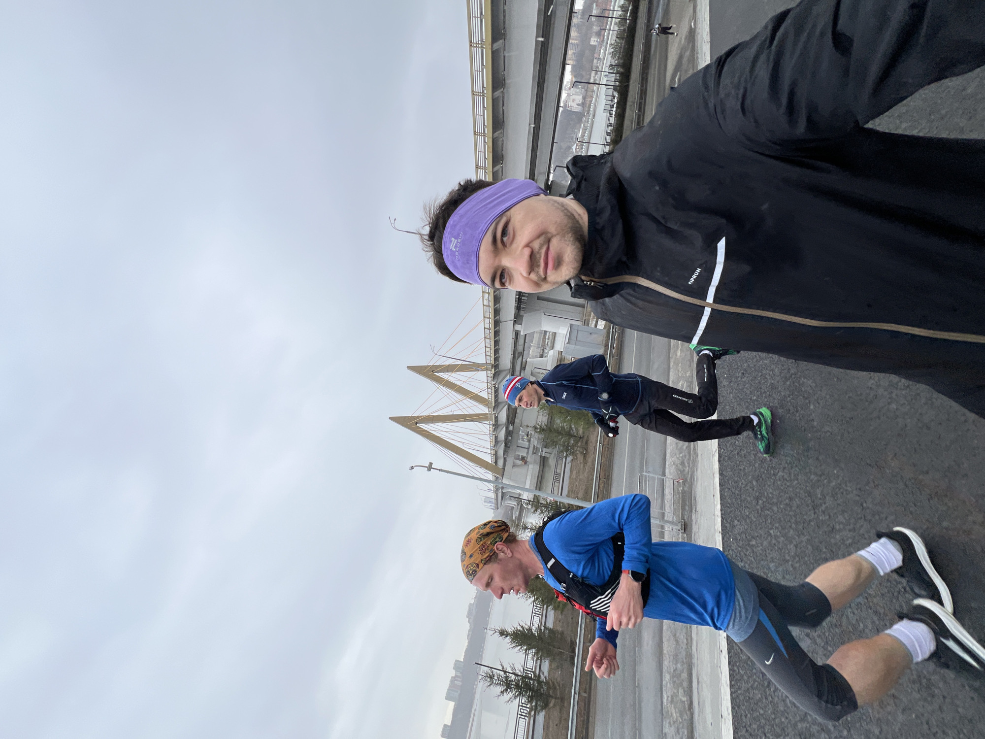 The image shows three people running in Kazan, Russia. The man in the foreground is taking a selfie. He is wearing a purple headband and a black running jacket. Behind him, two other runners are visible. One is wearing a blue long-sleeved shirt and black running tights with white socks and black and white running shoes, and has a patterned head wrap. The other runner is wearing a dark blue jacket, black running tights, and green running shoes. In the background is a grey sky, a river, and buildings including the Millennium Bridge, identified by its orange cables. The foreground is asphalt.