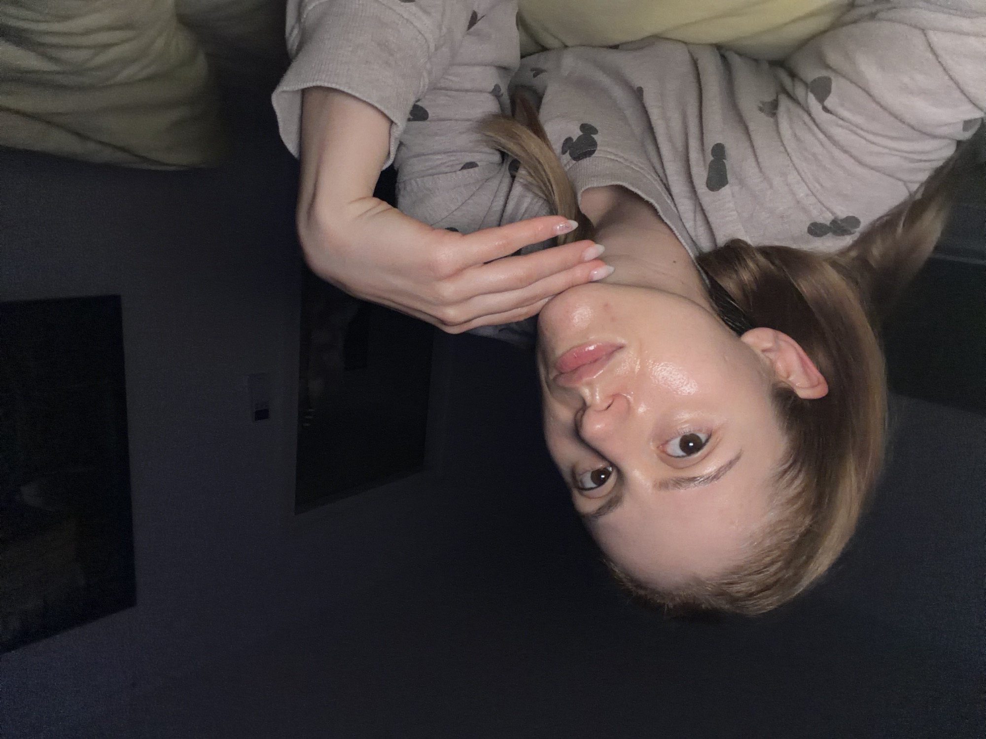 A medium shot shows a light-skinned woman with blonde hair and brown eyes lying in bed. She is wearing a grey long-sleeved shirt with a pattern of small grey mice. Her right hand is resting on her chin, and her left arm is bent at the elbow and visible to the left of her face. The woman's face is illuminated, and her skin appears to be shiny and possibly wet. The background is dark, with a hint of a wall and what appears to be a dark picture frame on the right side. The image appears to be taken in a bedroom at night. There is no discernible location in the city of Helmond, Netherlands.