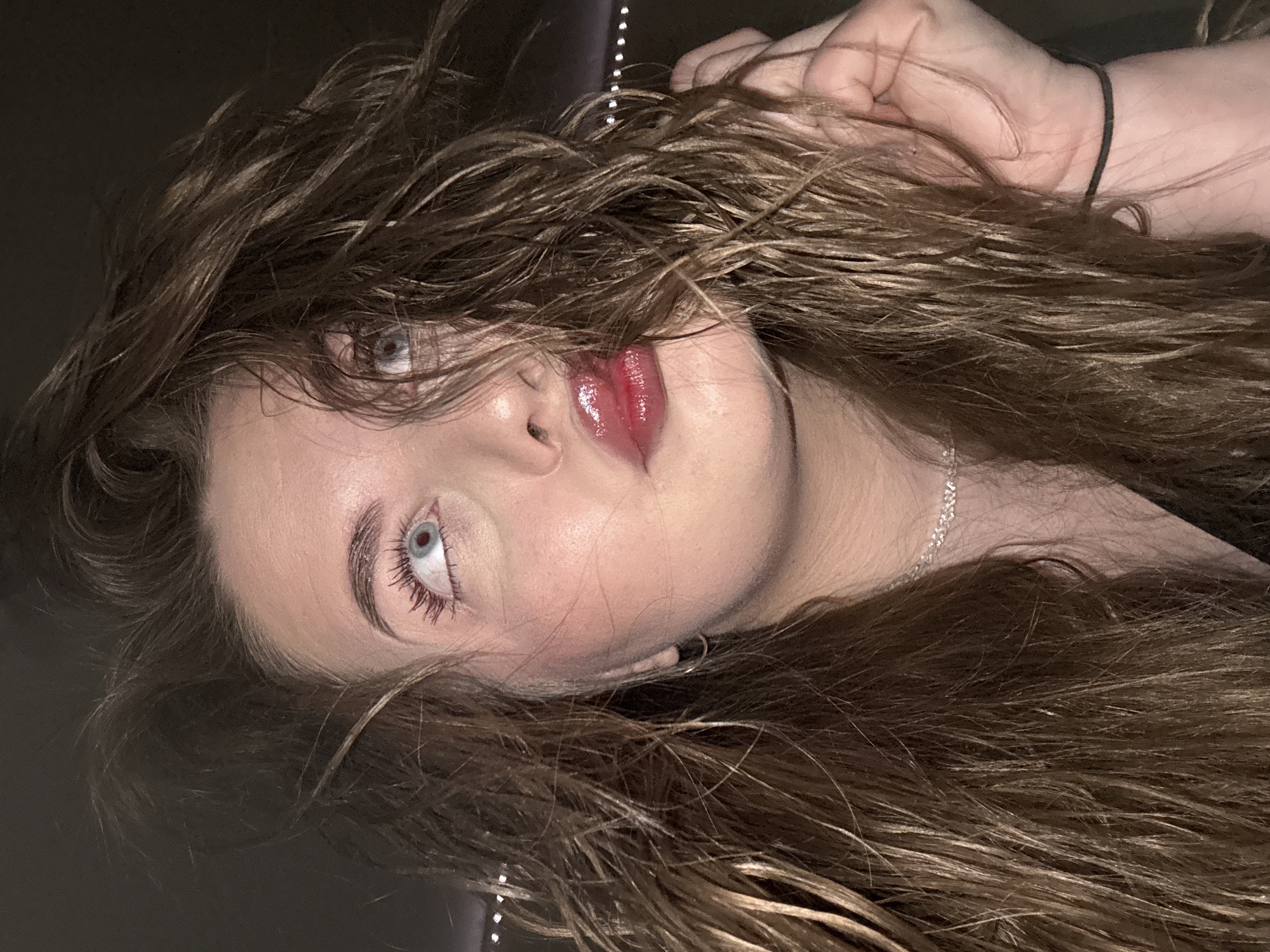 A young woman with light skin, light blue eyes, and brown, wet-looking hair is the subject of the image. She is angled to the left, with her face appearing to look up and slightly to the left. The woman has full lips with a glossy, red lipstick. Her hair is partially obscuring her face, and a silver necklace is visible around her neck. Her hand is near her hair at the top of the frame. The background is dark and indistinct. There is no indication that the image was taken in Chaikovsky, Russia.