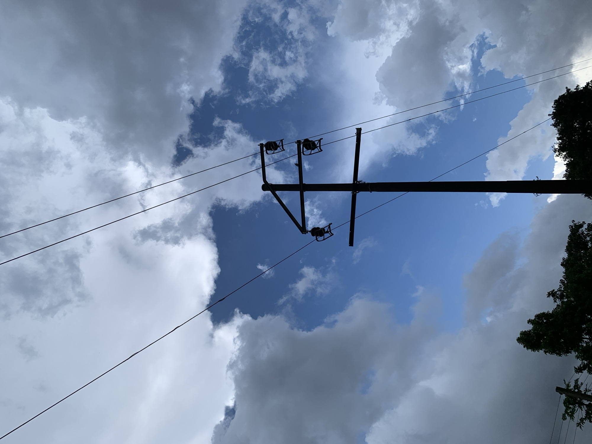 This media file depicts a scene with a prominent utility pole and wires against a dramatic sky filled with clouds. The sky transitions from a bright blue in some areas to dark, heavy storm clouds in others, suggesting dynamic weather. The utility pole is silhouetted against the sky, with visible insulators and wires stretching across the frame. A portion of a dark green tree is visible on the right side of the image, its branches reaching towards the sky. The perspective is looking upwards, emphasizing the vastness of the sky and the scale of the utility infrastructure. The lighting suggests it is daytime, with the sun's position creating strong contrasts between the illuminated and shadowed parts of the clouds. There are no people or explicit text visible in the image. The scene evokes a sense of the outdoors, possibly in a rural or semi-rural setting in Kuru Karama, Nigeria, with the weather contributing to a somewhat imposing or dramatic atmosphere.