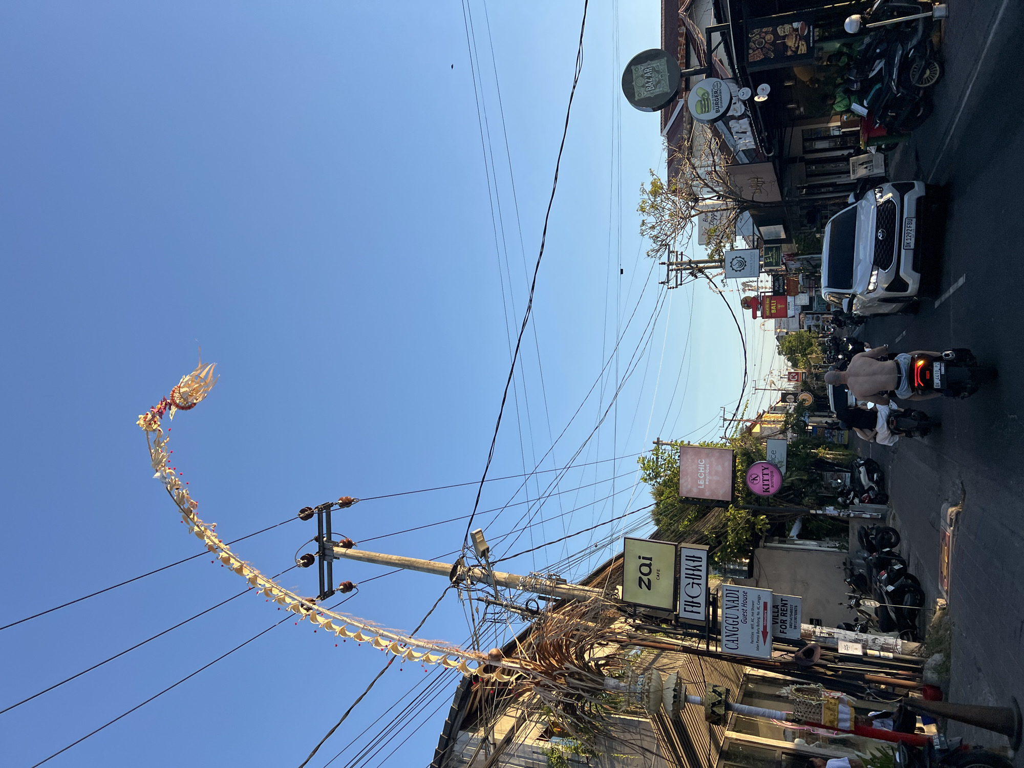 The image depicts a street scene in North Kuta, Indonesia, likely during the late afternoon. The sky is a clear, bright blue, with several power lines crisscrossing overhead. A decorative, curved banner, possibly a Penjor, arches over the street, suspended from a utility pole adorned with ceramic insulators. The banner appears to be made of woven straw and decorated with red and white elements.
On the left side of the street, a building with a weathered facade is visible, featuring architectural details and some greenery. Several signs are attached to a utility pole, including one for "zai CAFE," "FUGUKU," "CANGGU NADI Guest House," and "VILLA FOR RENT." Further down the street, more signs are visible for businesses such as "LE CHIC BOUTIQUE," "KITTY BOUTIQUE," and "BAZAR," which appears to be a restaurant with images of food.
The street is paved and has white lane markings. Several motorbikes are parked along the side of the road, and others are in motion. Two people are riding on a motorbike in the foreground, with the driver shirtless. A white SUV is also driving down the street, with its headlights on. The overall impression is a bustling street in a tropical urban environment, likely a popular tourist area given the presence of guest houses and boutiques. The lighting suggests the sun is low in the sky, casting shadows.