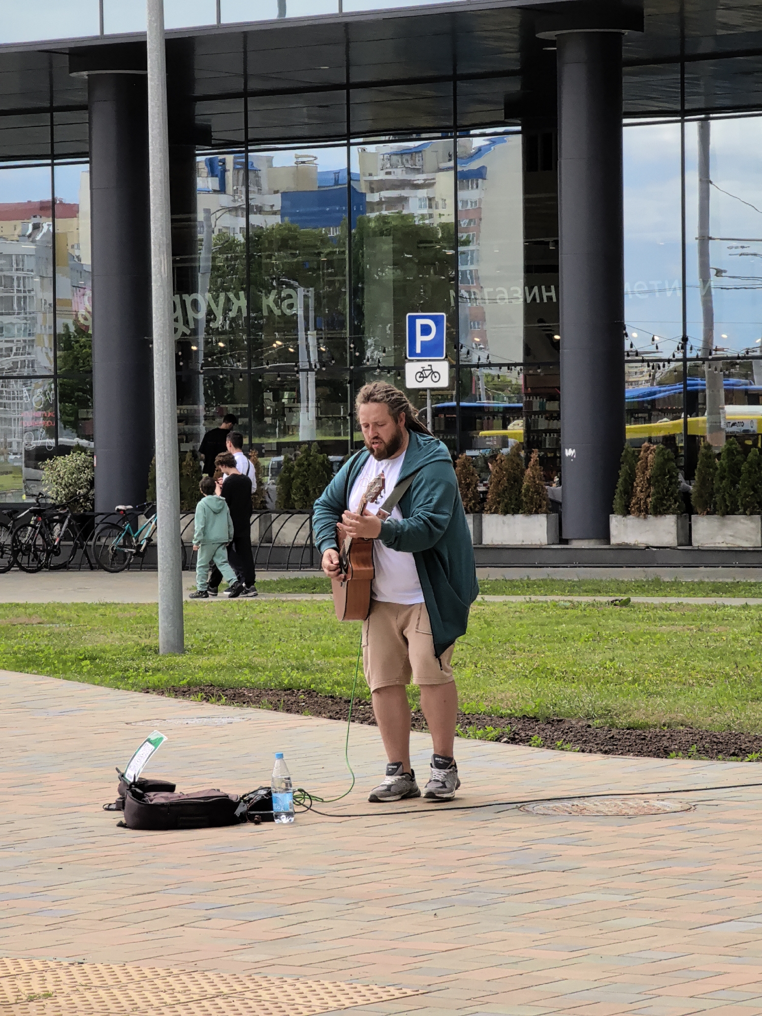 A male street performer with dreadlocks plays an acoustic guitar while standing on a paved area in front of a building with large glass windows. He is wearing a teal hoodie over a white t-shirt and beige shorts, along with grey athletic shoes. In front of him, on the pavement, is a dark-colored bag, a water bottle, and what appears to be a small speaker or amplifier with a green wire. Behind him, several people are walking, including a child in a light blue tracksuit. The building behind them reflects trees, other buildings, and the sky. There are planters with small evergreen trees in front of the building. Bicycles are parked on the left side of the image. The scene appears to be in an urban setting, possibly a plaza or pedestrian area.