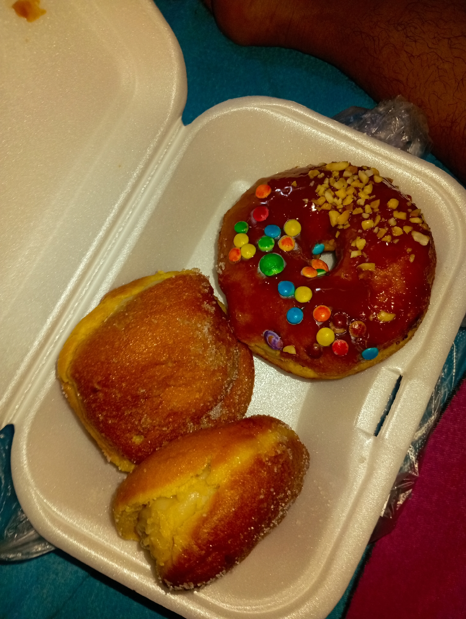 A white styrofoam container holds a donut with a shiny red glaze, colorful candies, and chopped nuts, along with two sugar-coated pastries. The container sits on a surface with teal and pink sections. A person's arm is visible in the background. The image was likely taken in Altagracia, Venezuela.