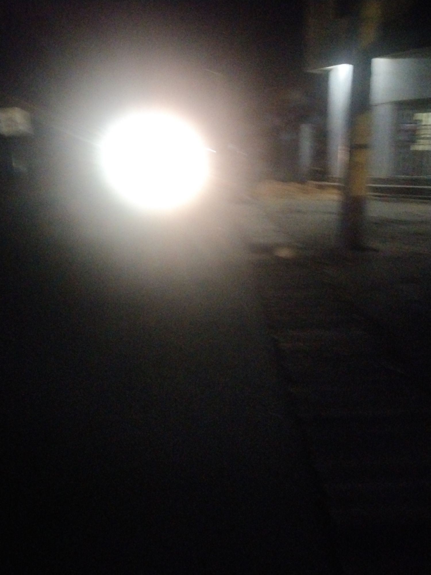 The image shows a night scene in Uyo, Nigeria. A bright light source dominates the left side of the frame, likely the headlight of a vehicle. The surrounding area is dark, with some streetlights visible in the background near a building, and a pole on the right of the frame. A road surface is visible near the bottom of the frame. The image is blurry.
