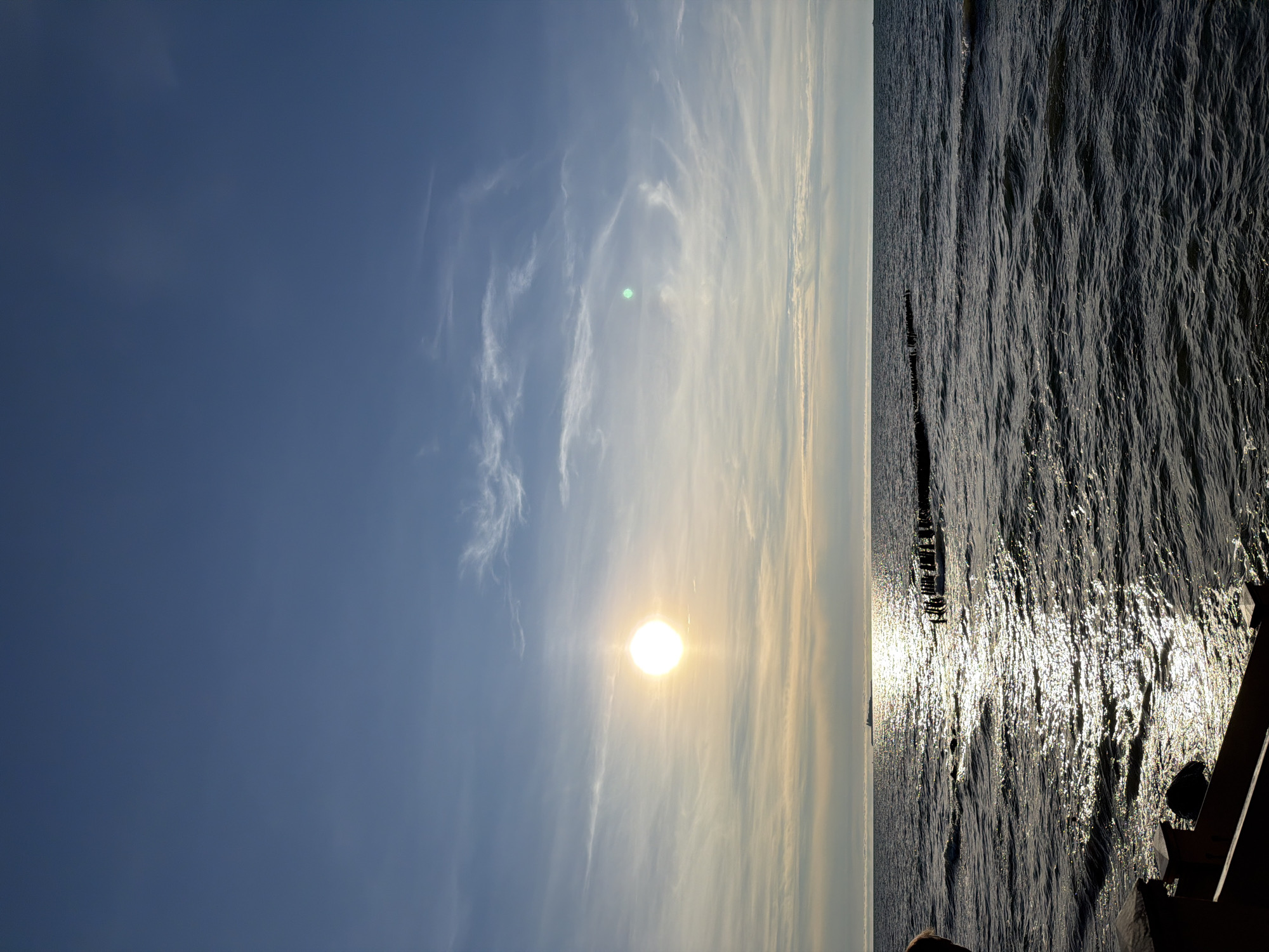 A wide shot captures a sunset over the Baltic Sea in Zelenogradsk, Russia. The sun, a bright orb, is low on the horizon, casting a shimmering path of light across the rippling water. A faint silhouette of a ship is visible on the horizon, to the left of the sun. In the foreground, a dark wooden railing frames the view, with a partial view of a person's head and shoulder on the far left. A line of weathered wooden posts emerges from the water in the mid-ground, suggesting remnants of a pier or breakwater. The sky is a gradient of blue and pale yellow, with wispy clouds stretching across. The overall scene conveys a sense of calm and natural beauty.