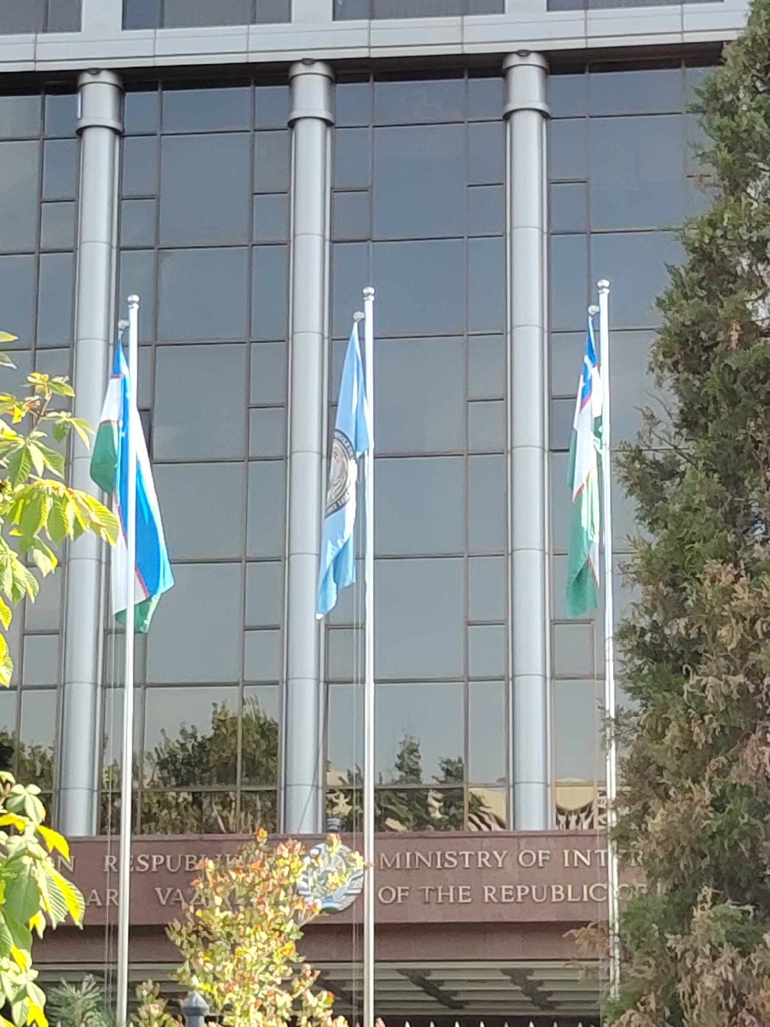 The image shows the Ministry of Foreign Affairs of Uzbekistan in Tashkent. The building is modern, with a glass facade and prominent silver columns. Several flags are flying in front of the building, including the flag of Uzbekistan and what appears to be a United Nations flag. The building has text inscribed on it that reads "MINISTRY OF INT R OF THE REPUBLIC OF". The reflection in the windows shows trees and the sky. There are green leaves in the foreground, suggesting it is spring or summer. The overall scene is well-lit, indicating it was taken during the daytime.