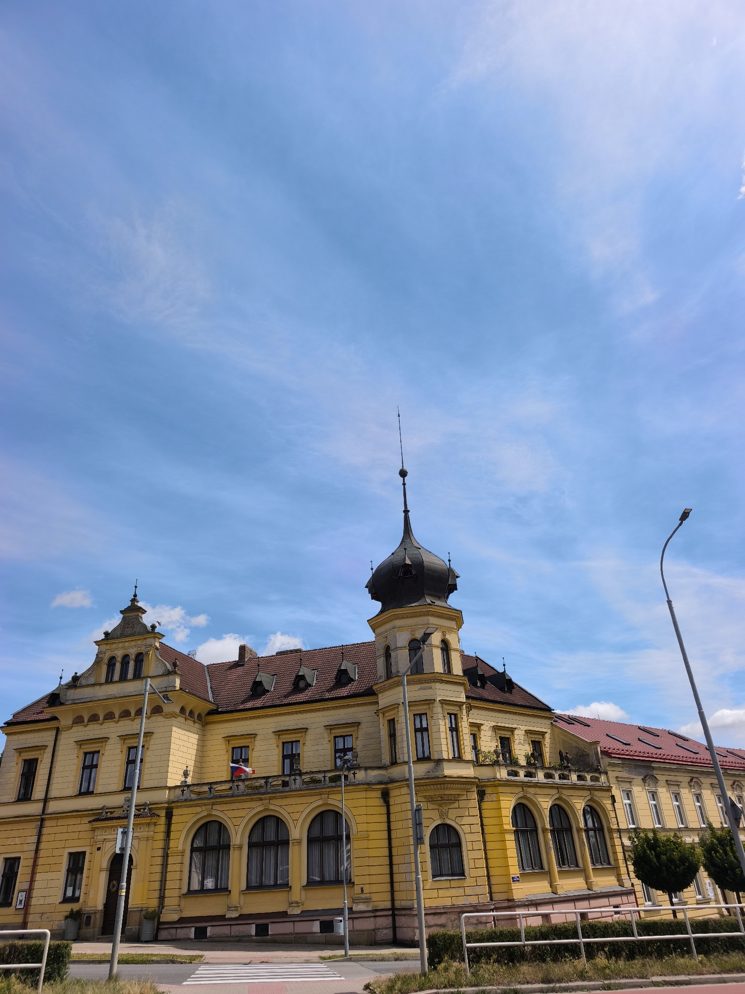 The image shows a bright yellow building with a red-tiled roof and a prominent black dome tower, located in the city of Vysoké Mýto, Czechia. The building features classical architectural elements, including arched windows, decorative cornices, and multiple stories. A flag, appearing to be the flag of the Czech Republic, is visible on a balcony. The sky above is a clear blue with wispy white clouds. In the foreground, there is a street with a pedestrian crossing, a lamppost, and some greenery. The scene appears to be a daytime view of a public building within an urban setting.