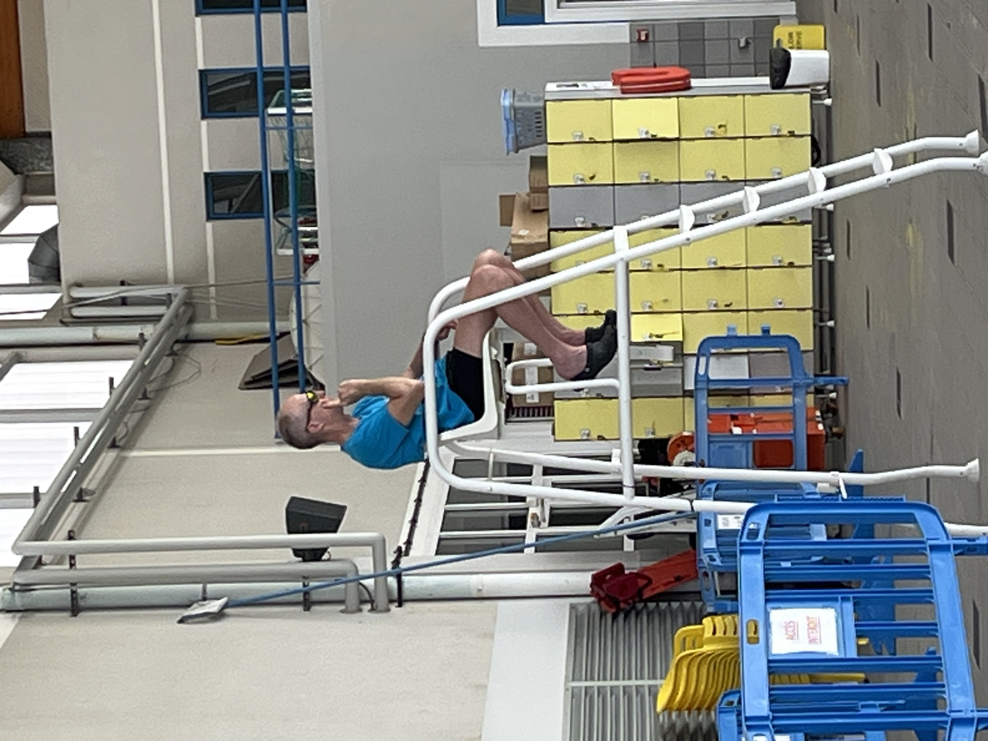 A man is seen on the side of a pool. The man is wearing black slip-on shoes, black shorts, and a short-sleeved blue shirt. The scene is located in Andrézieux-Bouthéon, France. Behind the man are white metal bars, yellow and gray lockers, and blue and yellow plastic chairs. A long, white horizontal pole runs behind the man as well.