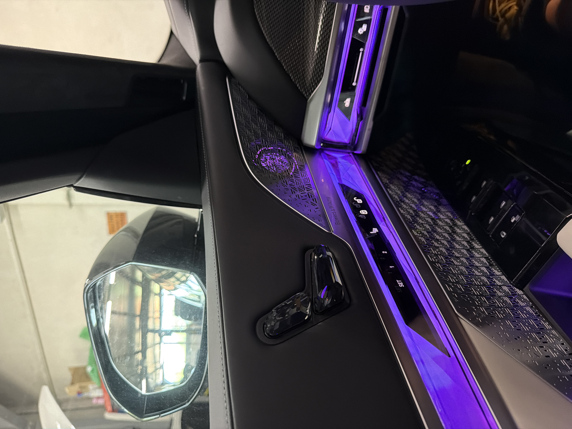 The image shows the interior of a luxury car, specifically the driver's side door panel, with ambient lighting. The door panel is black leather with chrome accents and features several buttons, including seat adjustment controls and controls for various car functions, such as locking doors and climate control. The ambient lighting is purple and illuminates the lower part of the door panel. The side mirror of the car is also visible, reflecting the interior of a garage. The car appears to be parked indoors. There are no people or animals visible in the image. The environment suggests a car repair shop or a showroom. The image is taken from a close-up perspective, focusing on the details of the car's interior. There is no information in the image that indicates the location of Burgas, Bulgaria.