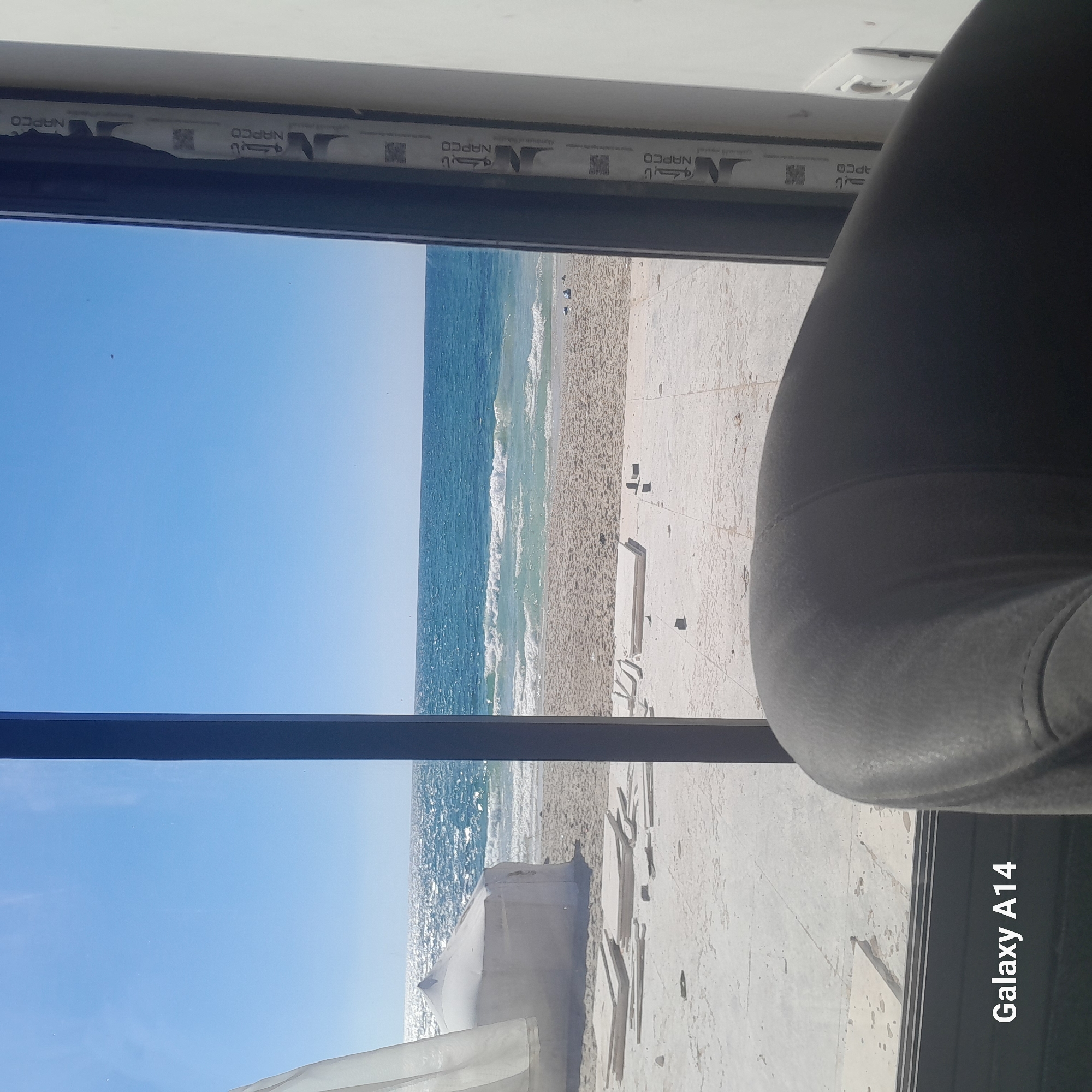 This media file is a photograph taken from inside looking out a window at a beach scene. The foreground features a dark, cushioned object, possibly a chair or sofa, taking up the right side of the frame. The window itself has a black frame, and the top part of the frame has a strip with repeating text and logos. The text appears to say "NAPCO" in a repeating pattern.
Beyond the window, the scene depicts a bright, sunny beach. The ocean is a vibrant blue, with gentle waves breaking on the shore. The sand is light in color and appears to be somewhat textured. There are a few dark specks on the sand that could be small objects or figures. In the middle distance, there's a white, tent-like structure on the beach. The overall impression is a clear, possibly daytime scene with good weather.
A strip of text in the bottom right corner reads "Galaxy A14". The image is oriented vertically, suggesting it was taken with a phone. The lighting and clear blue sky indicate a sunny day, likely during daylight hours. The overall mood is relaxed and serene, consistent with a beach view.