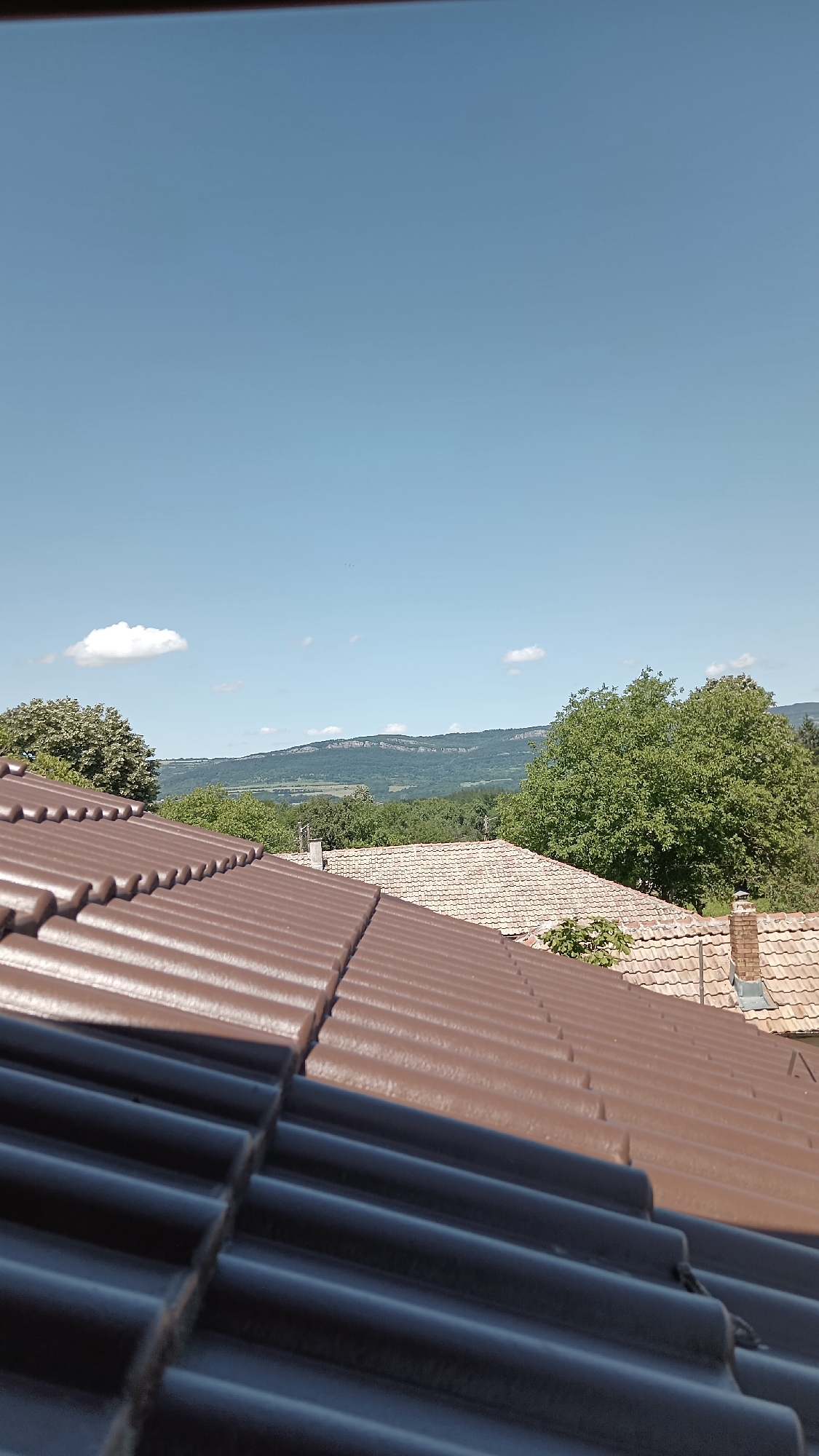 A high-angle, outdoor, medium shot captures a clear blue sky over a rural landscape of rolling hills and vegetation. In the foreground, two tiled roofs dominate the lower portion of the frame. The roof closest to the viewer is dark, almost black, and features rows of ribbed tiles. Behind it, another roof made of reddish-brown ribbed tiles is visible, with a portion of a lighter-colored, tiled roof and a brick chimney extending upwards on the right. A distant, flat-topped mountain range forms the horizon line, partially obscured by scattered trees and the hazy atmosphere. A single, puffy white cloud drifts in the upper left quadrant of the sky. The image appears to be a view from a rooftop, looking out over the village of Mihaylovtsi, Bulgaria.
