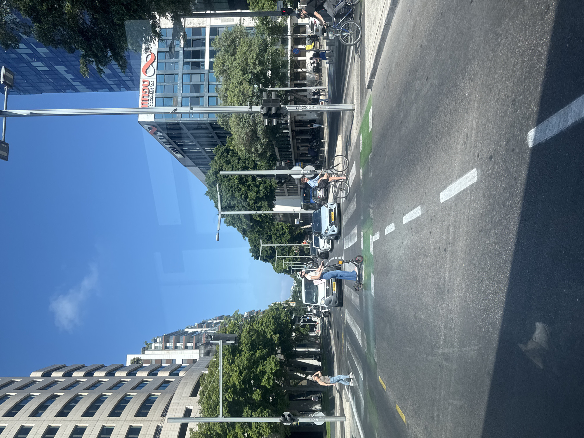 This is a street-level, eye-level view of a city intersection in Tel Aviv, Israel, on a clear, sunny day. The perspective appears to be from a vehicle. The road has distinct white and yellow lane markings, and a green-painted bike lane is visible across the intersection.
In the foreground, a woman on an electric scooter is crossing the street, wearing a helmet and white top with blue jeans. To her left, another woman is walking across the pedestrian crossing, wearing a beige tank top and blue jeans.
Further into the intersection, several vehicles are present, including a white van, a white sedan, and other cars. On the right side of the image, two men are riding bicycles across the intersection. One is on a dark-colored bike and the other on a blue bicycle.
The background features tall buildings with modern architecture. On the right, a building with a prominent red and white logo that reads "Tefen" is visible, suggesting this area is likely a business district. Trees line the sides of the street, providing shade. Traffic lights are visible at the intersection. The sky is a bright, cloudless blue.
