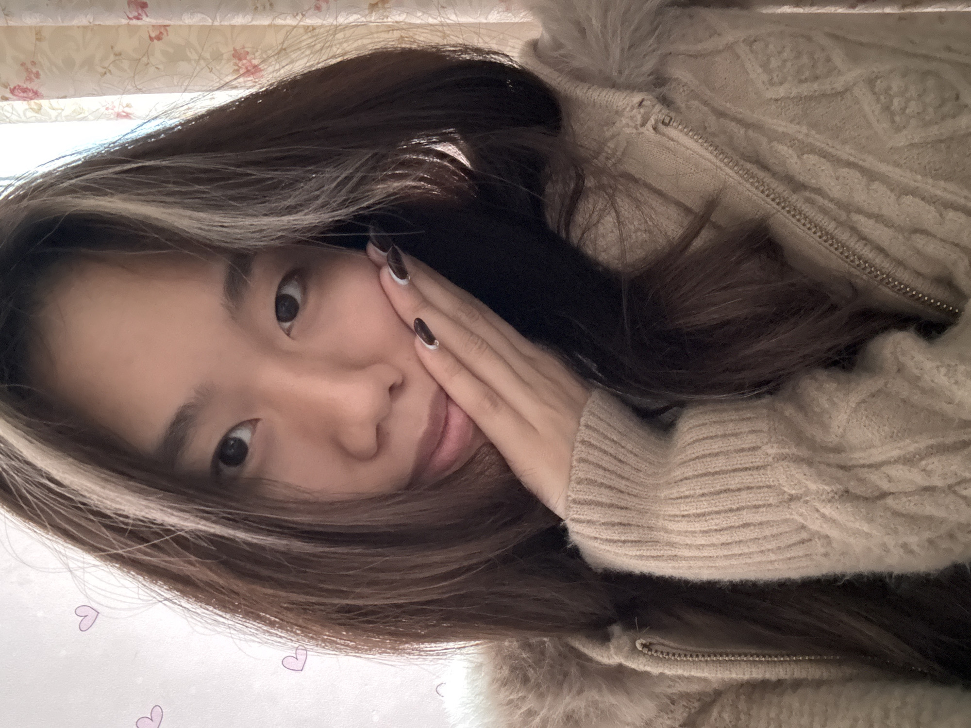 A young woman is facing the camera in an indoor setting. She has long, dark hair with a single lighter highlight near her forehead, and wears a light-colored, cable-knit sweater with fur trim. Her left hand is lightly touching her face, and her nails are painted with a dark color and a white design. The background is out of focus but appears to be a light-colored wall with a floral pattern at the top and small, light purple hearts near the bottom. The image is rotated, with the left side of the image oriented towards the top. It is not possible to discern the location in Shapingba District, China, based on the visual information provided.