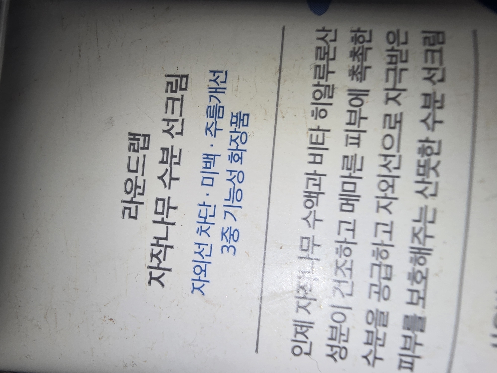 The image displays text in Korean. The text appears to be from a product label or description. The main heading is "라온드랩" (Raon Deu-raem), followed by "자작나무 수분 선크림" (Birch Water Sun Cream). Below this, there is "자외선 차단 · 미백 · 주름개선" (UV Protection · Whitening · Wrinkle Improvement) and "3중 기능성 화장품" (Triple Functional Cosmetic). The lower part of the text describes the product's benefits, stating it provides moisture and soothes dry, irritated skin with birch sap and hyaluronic acid, acting as a UV protection and moisture-rich sunscreen. There is no indication of the city of Ulan-Ude, Russia, in the image. The scene is a close-up of printed text on a textured surface.