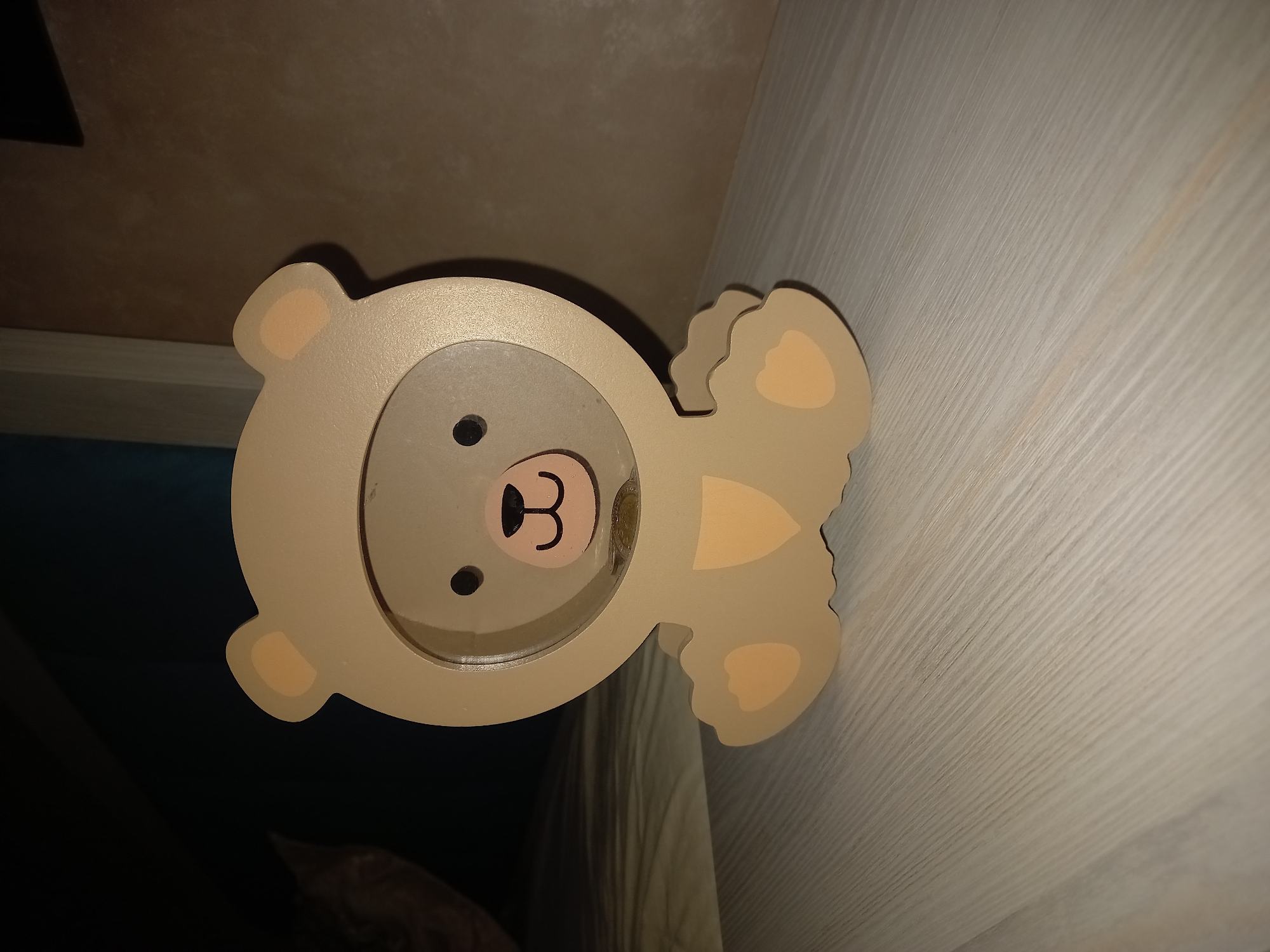 A wooden coin bank shaped like a cartoon bear is placed on a light-colored wooden surface. The bear is beige with lighter beige accents on its ears and paws. It has a round opening in the face area through which two coins are visible. The background is dark and out of focus, with a hint of what appears to be a bed or couch. There is no indication of the city of Tashkent, Uzbekistan.