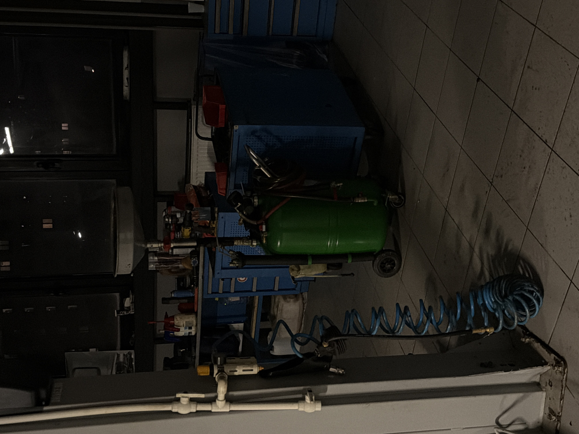 The image shows the interior of a garage or workshop. Dominating the scene is a blue metal tool cabinet, likely on wheels, filled with various tools and equipment. A green air compressor is positioned next to the cabinet, with a coiled blue air hose extending towards the foreground. The floor is tiled. The room is dimly lit, with light entering from a nearby window. Based on the window's reflection, it appears to be nighttime outside. The location in the city of Izhevsk, Russia is not evident.