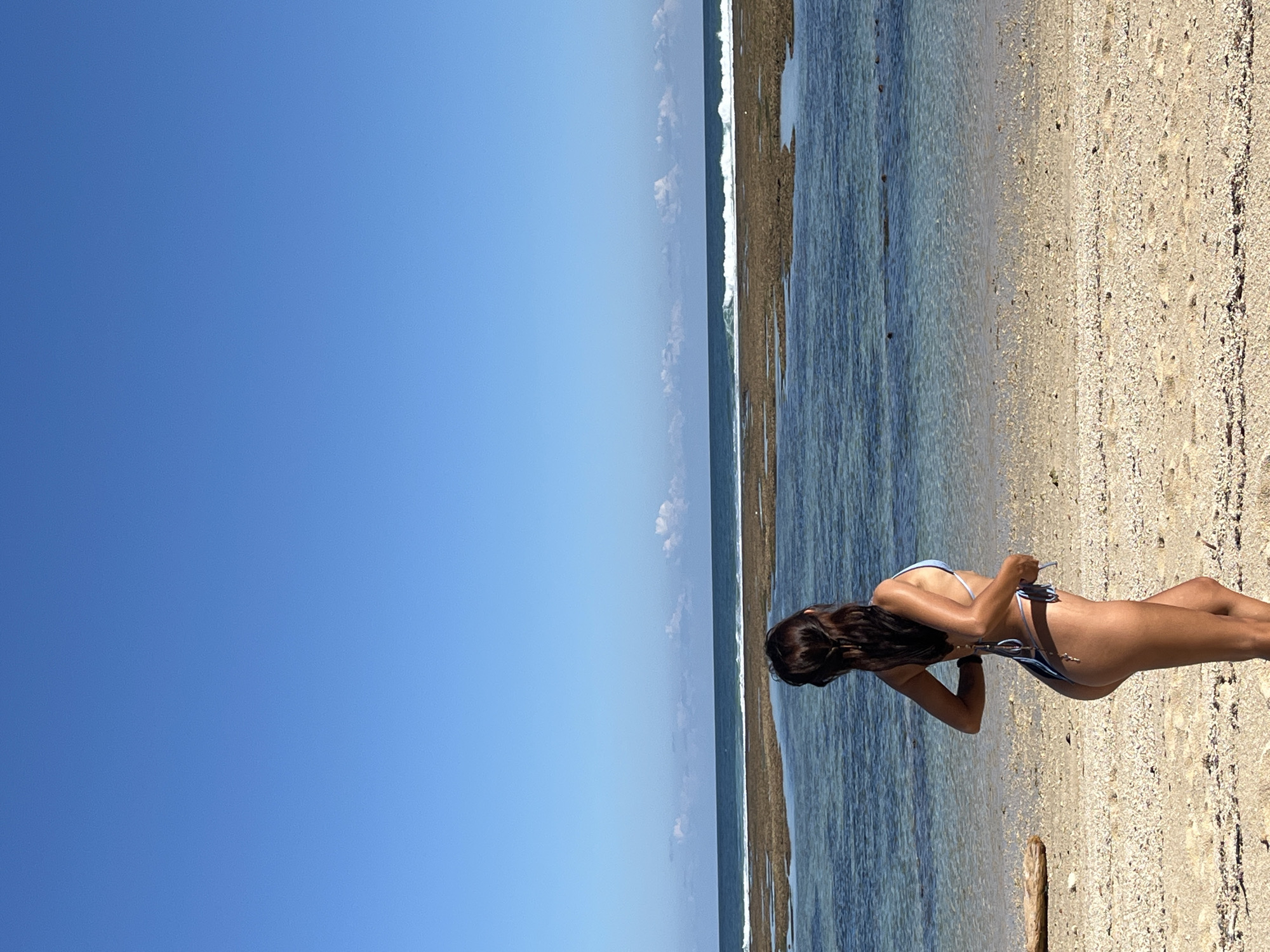 This image depicts a serene beach scene in Kuta Selatan, Indonesia, under a bright, clear blue sky. A person, appearing to be a woman, stands on the sandy shore, positioned towards the right side of the frame and viewed from behind. She is wearing a light-colored (possibly blue or white) bikini and has long, dark hair flowing down her back. Her hands are raised, seemingly adjusting the straps or ties of her bikini top.
The setting is characterized by the expansive, clear blue sky occupying the upper left portion of the image. The ocean, with its transparent, turquoise waters, stretches across the middle of the frame, showing a distinct line of breaking waves or a rocky shelf further out from the shore. The light-colored sandy beach, rich with tiny shells and pebbles, occupies the foreground and extends towards the water's edge. A small piece of driftwood is visible on the sand near the water. The strong, direct sunlight suggests it is daytime, likely mid-morning or early afternoon. No text is visible in the image.