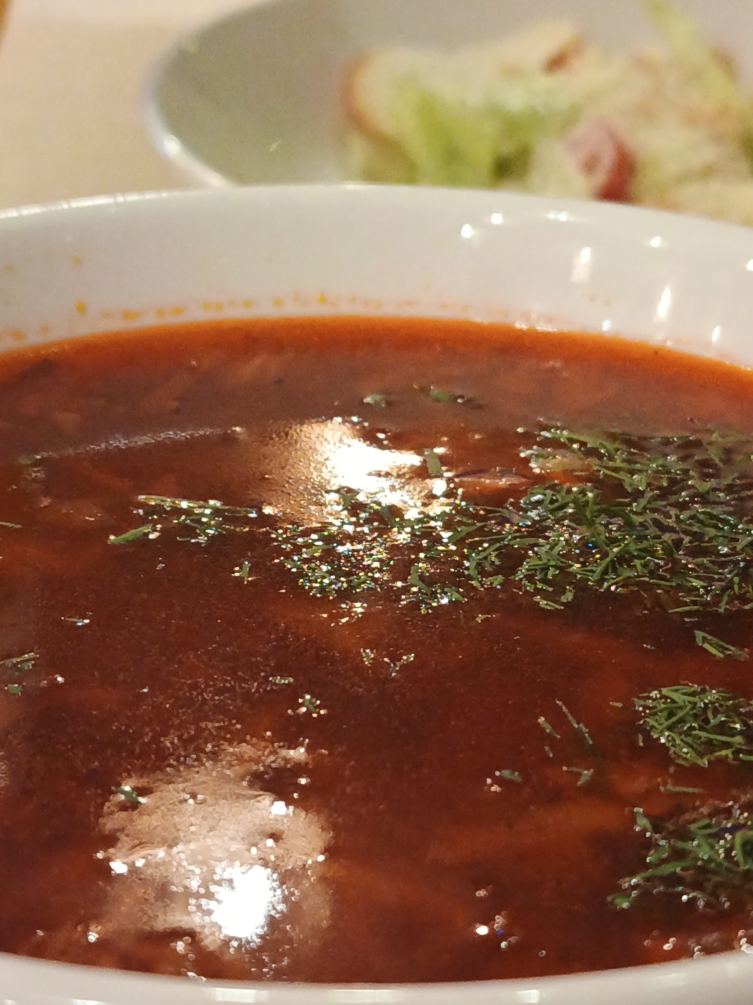 A close-up shot focuses on a bowl of rich, dark red soup, likely borscht, garnished with chopped green dill. The soup has a glistening surface, reflecting the light in small, bright spots. The bowl itself is white ceramic. In the blurred background, another bowl or plate is visible, containing what appears to be a salad with green leaves and chunks of food, possibly meat or croutons. The overall setting seems to be a restaurant or dining environment, given the presentation of the food and the tableware. The image does not provide any specific geographical markers to pinpoint a location within Bac Nha Trang Ward, Vietnam.