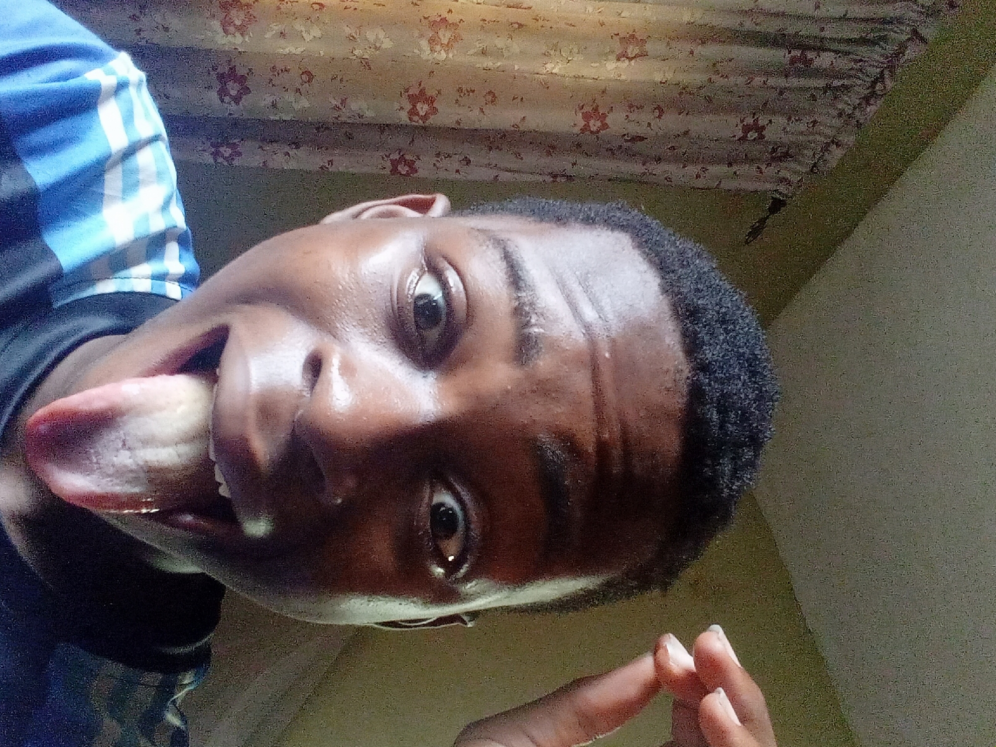 The image features a close-up, low-angle selfie of a young Black person taken in what appears to be an indoor setting in Nkpor, Nigeria. The person is looking directly at the camera with a playful expression, sticking out their tongue. Their right eye (viewer's left) is wide open, while the left eye is partially obscured. Their skin is shiny, possibly indicating sweat or a humid environment. They have short, dark, tightly curled hair.
The person is wearing a dark garment with prominent blue and white stripes visible on the shoulder area, resembling an athletic jersey or t-shirt. Part of a finger, likely from their left hand, is visible near their chin.
The background consists of two main elements: in the upper left, a floral curtain or fabric with a light base and reddish-brown floral patterns is visible. The rest of the background, primarily on the right and bottom, is a plain, light-colored wall, possibly green or beige, suggesting a simple room. The lighting appears to be natural, indicating it's daytime. The overall mood conveyed is lighthearted and expressive. No visible text is present in the image.