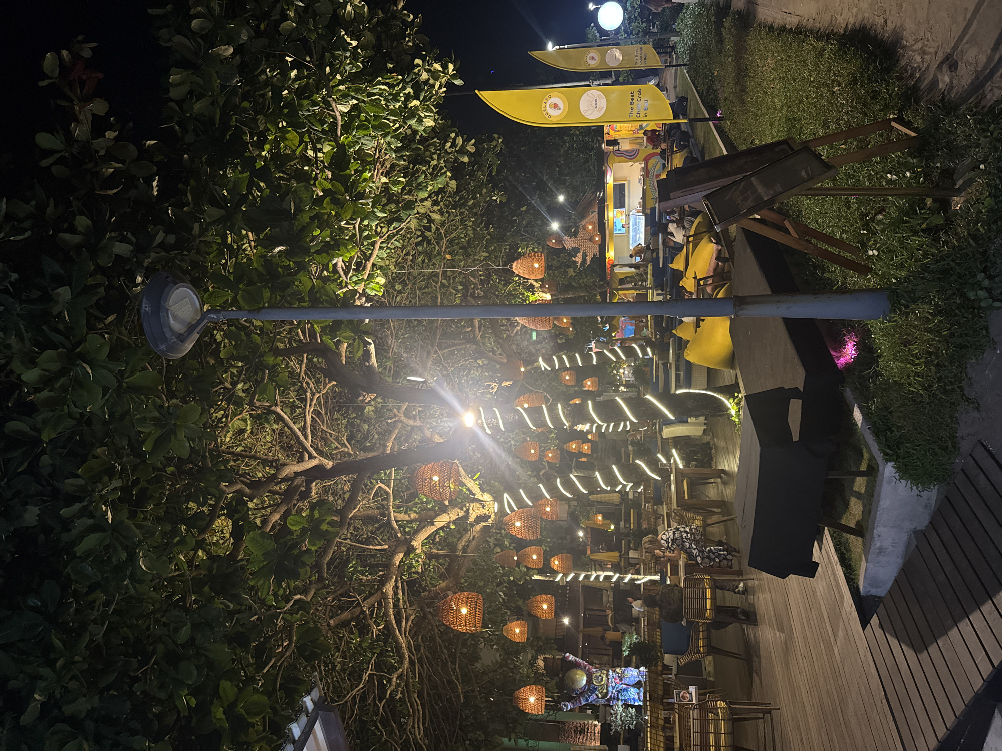 This image depicts an outdoor seating area at night in Kuta, Indonesia, likely a restaurant or cafe. The scene is illuminated by string lights wrapped around tree trunks and numerous woven pendant lamps hanging from branches. A tall lamppost stands near the foreground. In the background, a yellow food truck with the text "The Best Chili Crab in Bali" is visible, alongside two yellow flags bearing similar text. Several tables and chairs are arranged throughout the space. Some patrons are seated at tables, while others are standing or walking. A large, dark-covered object resembling a low table or bench is situated in the foreground. The ground is covered with wooden decking in the foreground, transitioning to grass and sand further back. The overall atmosphere is one of a lively evening gathering.