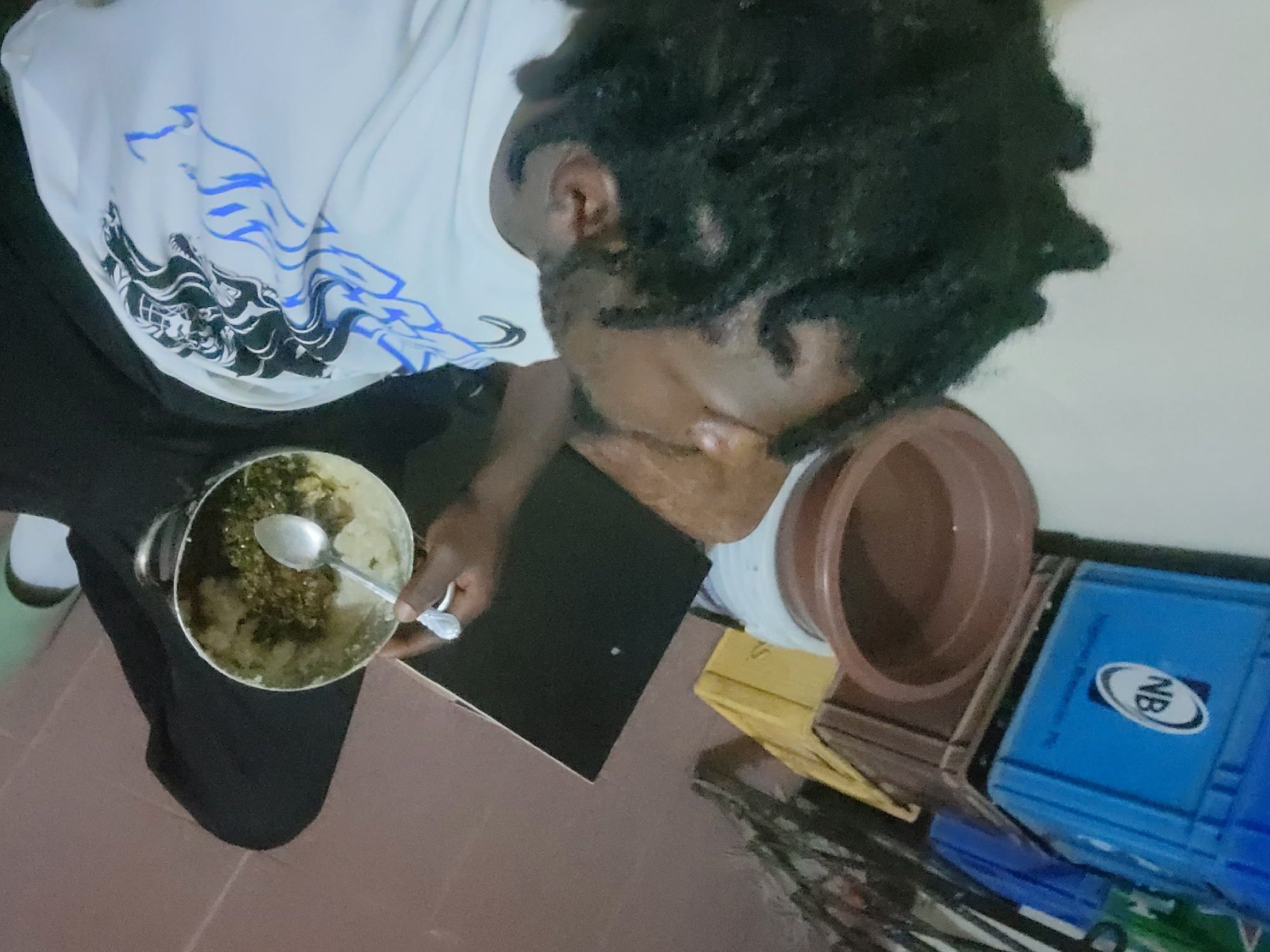 A person with dark skin and dreadlocks is shown from above, apparently eating from a metal pot filled with a light-colored food mixed with dark green vegetables. The person wears a white t-shirt with black and blue designs on the front and black trousers. The person sits at a black table that sits on top of a brownish cardboard, close to a brown vase and a blue NB-labeled plastic container. The background appears to be an indoor space with off-white walls. Although the location cannot be definitively verified as Jos, Nigeria based solely on the visual information, there are no visual elements contradicting it.