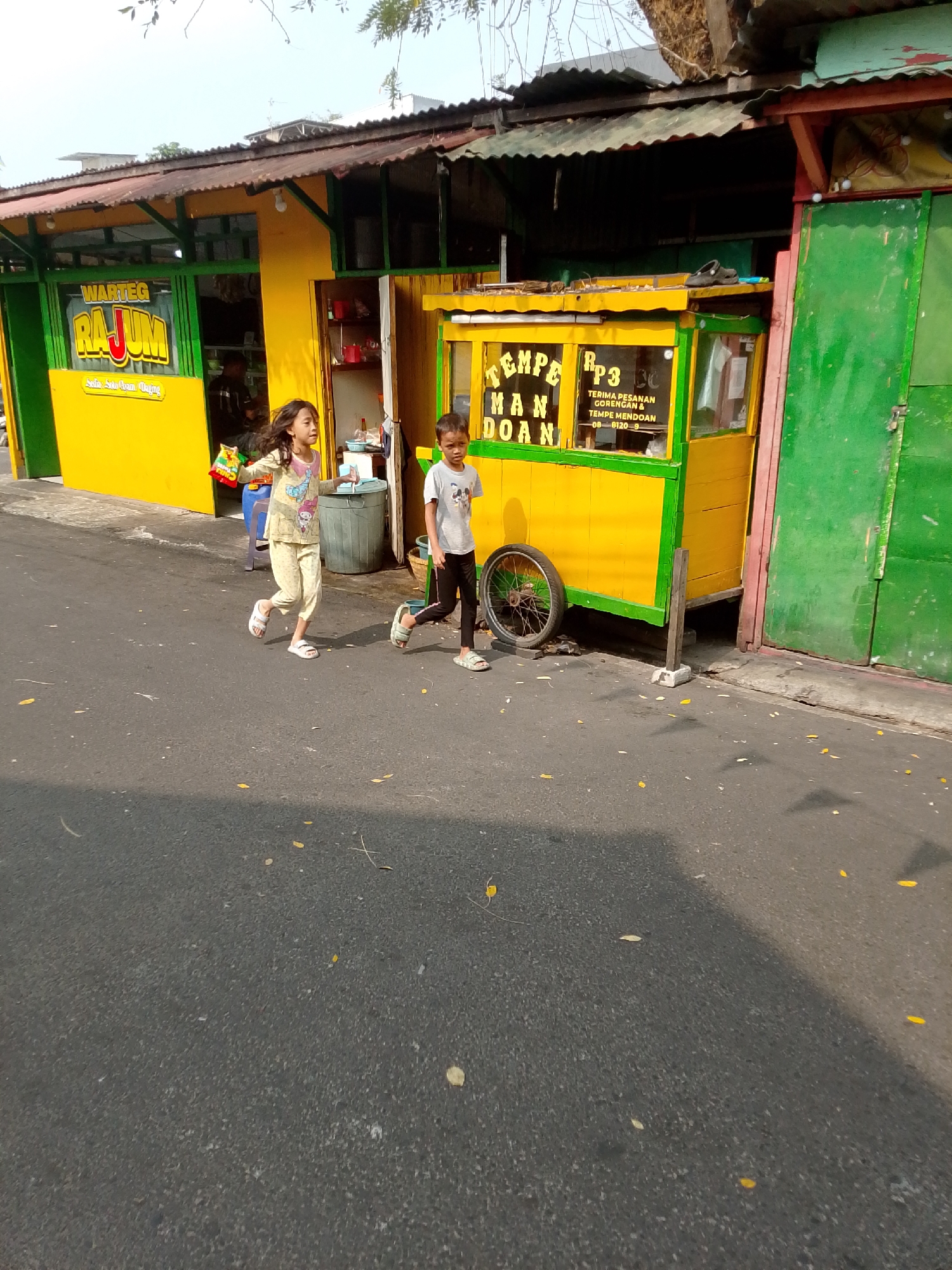Two children are running on an asphalt road in front of a food stall. The stall is brightly colored yellow and green, with signage indicating it sells "TEMPE MAN DOAN" and "WARTEG RAJUM". The child on the left, a girl, is wearing pajamas and appears to be holding a snack bag. The child on the right, a boy, is wearing a t-shirt and dark pants. The setting appears to be a street in East Jakarta, Indonesia, with a corrugated metal roof visible over the stall and trees in the background. The sun is casting shadows on the road.