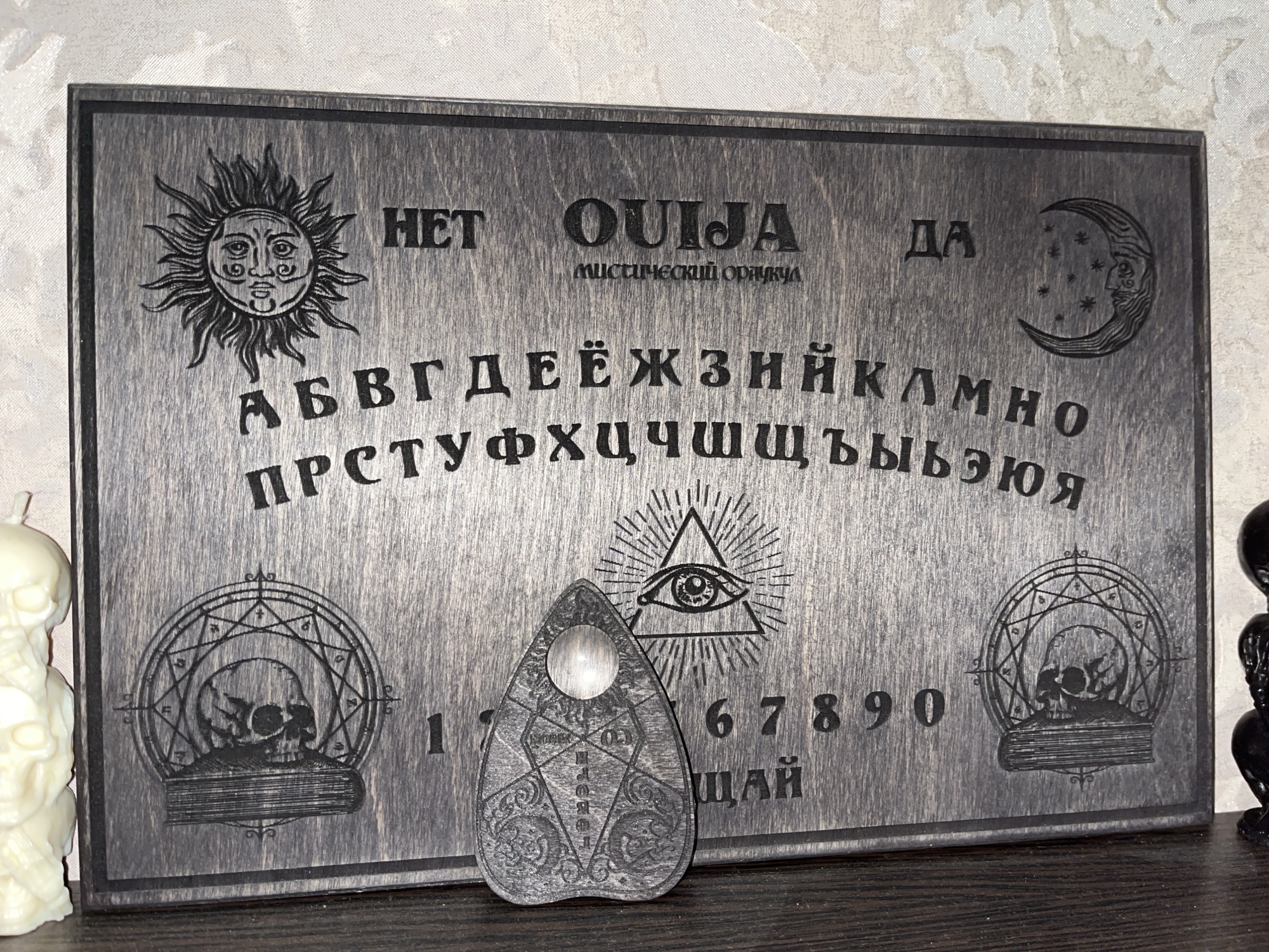 The image shows a Ouija board made of dark wood. The board has various symbols and letters etched into it, including the sun, moon, stars, skulls, and an all-seeing eye. There are also Cyrillic letters and numbers present. A heart-shaped planchette with an eye symbol is placed on the board. The Ouija board is resting on a dark wooden surface. To the left of the board, a light-colored skull-shaped candle is partially visible. To the right, a dark figure, possibly another decorative object, is partially visible. The background is a textured, light-colored surface. There is no visible indication of the city of Bryansk, Russia, or any specific location.