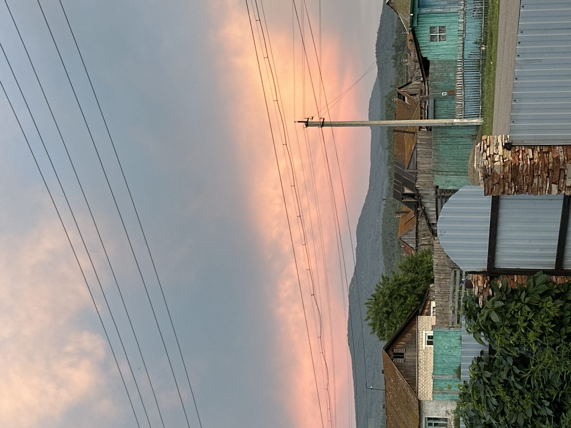 In the city of Unknown, Unknown, a rustic village scene unfolds under a twilight sky painted with hues of pink and gray. Overhead, a network of power lines crisscrosses the frame, stark against the softened light. In the middle ground, a cluster of weathered wooden houses and fences, some painted a vibrant teal, form a humble village. A dirt road meanders through the settlement. In the background, a range of soft, green mountains creates a picturesque backdrop. The foreground is occupied by a metal fence and lush green foliage, hinting at the encroaching natural world. The overall impression is one of quietude and rural charm, a peaceful snapshot of a life lived close to nature.
