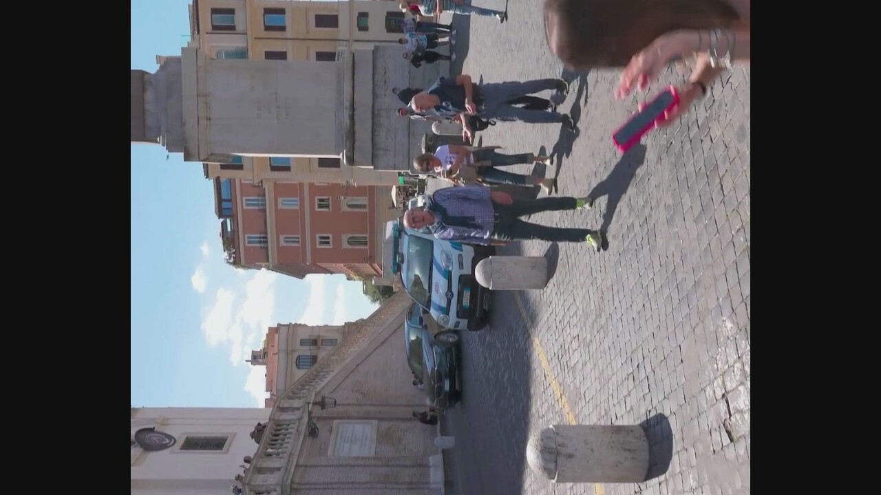 In a sunlit, cobblestone square in Nepi, Italy, a man wearing a blue plaid shirt and a dark sweater draped over his shoulders stands facing the camera. He has glasses and is wearing dark pants and light green sneakers. To his left, a dark sedan is parked near a white and blue police car. Behind the man, a woman with blonde hair is walking away from the camera, carrying a beige bag. To the right of the blonde woman, a man in a dark blue shirt and dark pants appears to be speaking with her, gesturing with his hands. In the background, several buildings with colorful facades and balconies are visible under a bright, partly cloudy sky. Several other people are scattered throughout the square. In the foreground, a woman with long brown hair is partially visible, holding a pink phone.
