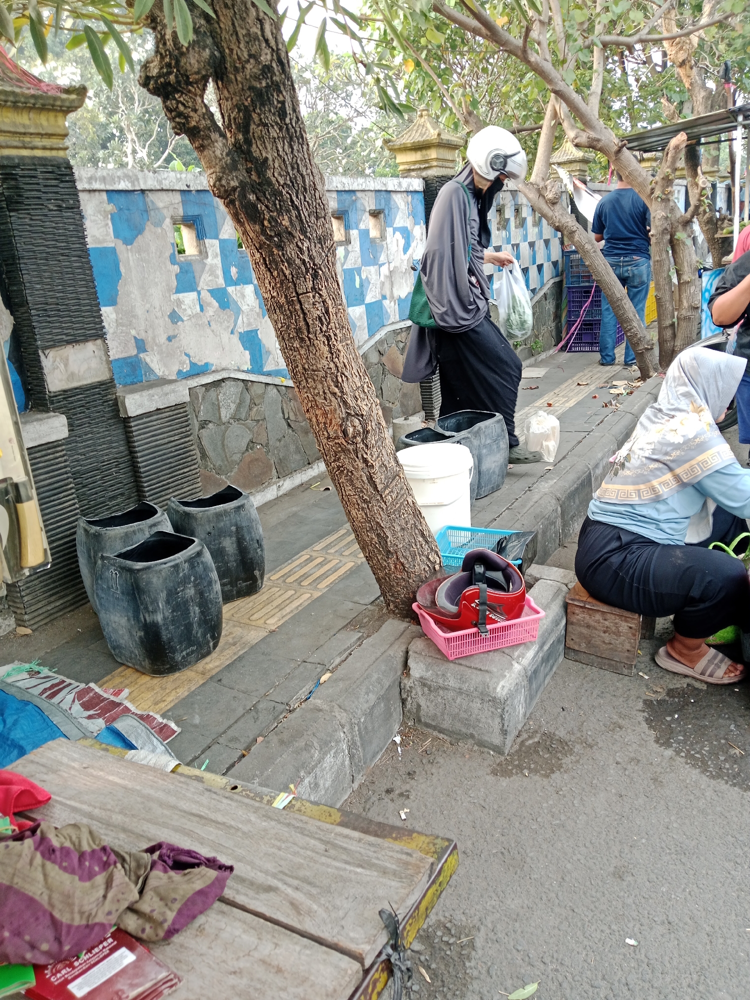 The image depicts an outdoor scene, likely a street or market area in Kediri City, Indonesia. In the foreground, a wooden table is partially visible, adorned with folded cloth items. To the left, three large, black, cylindrical containers are placed on a sidewalk. A tree trunk, with textured bark, stands prominently in the mid-ground, leaning towards the right side of the frame. Behind the tree, a woman wearing a grey hijab and a white helmet walks, carrying a white plastic bag. Further in the background, a man in a blue t-shirt and jeans stands near a stack of colorful crates. Another person, wearing a light blue hijab and black pants, is seated on a wooden stool on the right side of the image, facing away from the camera. The environment shows a tiled wall with a blue and white checkered pattern, and some parts of the wall appear weathered. The ground is paved with a combination of concrete and asphalt, with visible yellow tactile paving on the sidewalk.