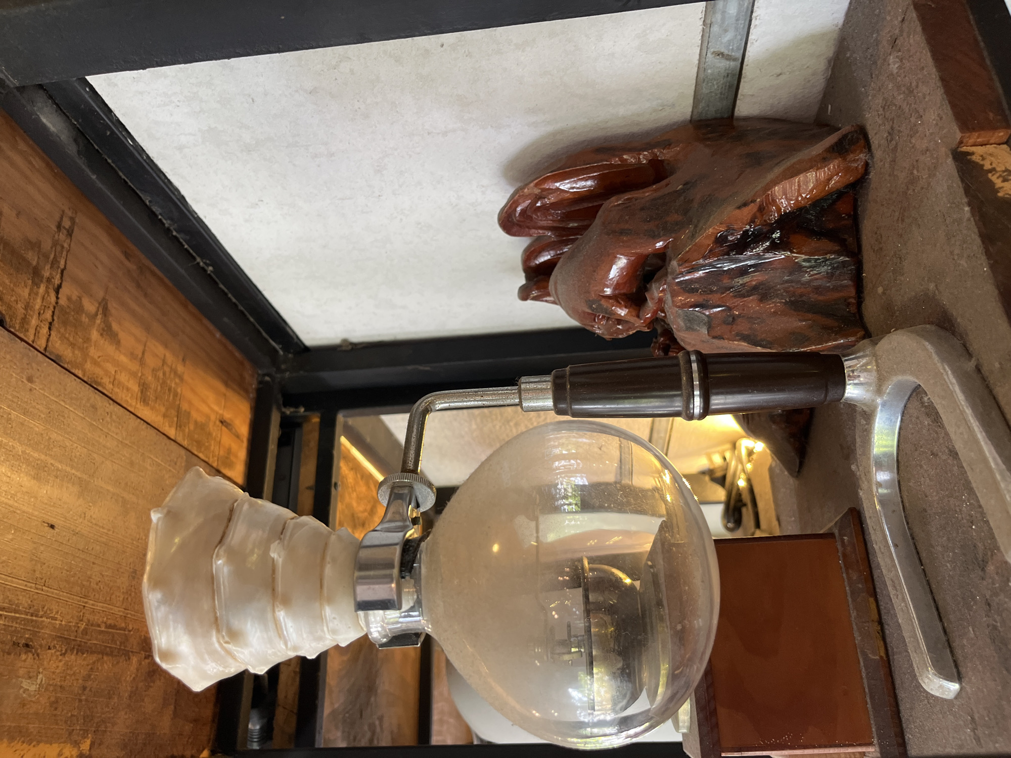 The image, oriented with the top toward the left, depicts a vintage coffee-making apparatus mounted within a structure that appears to be part of a building or display, possibly a coffee shop in Bac Nha Trang Ward, Vietnam.
The coffee maker has a glass bulb, somewhat dusty, and a side handle with a brown grip. The glass connects to a ridged white filter. It is attached to a metal stand on the right side.
Above the coffee maker, on what would be the ceiling or top portion of the structure, there's a glossy, dark wood sculpture and a white surface, potentially a ceiling tile. The structure itself seems to be constructed of a combination of dark metal framing and wood paneling. The perspective is an upward view, likely taken from below.